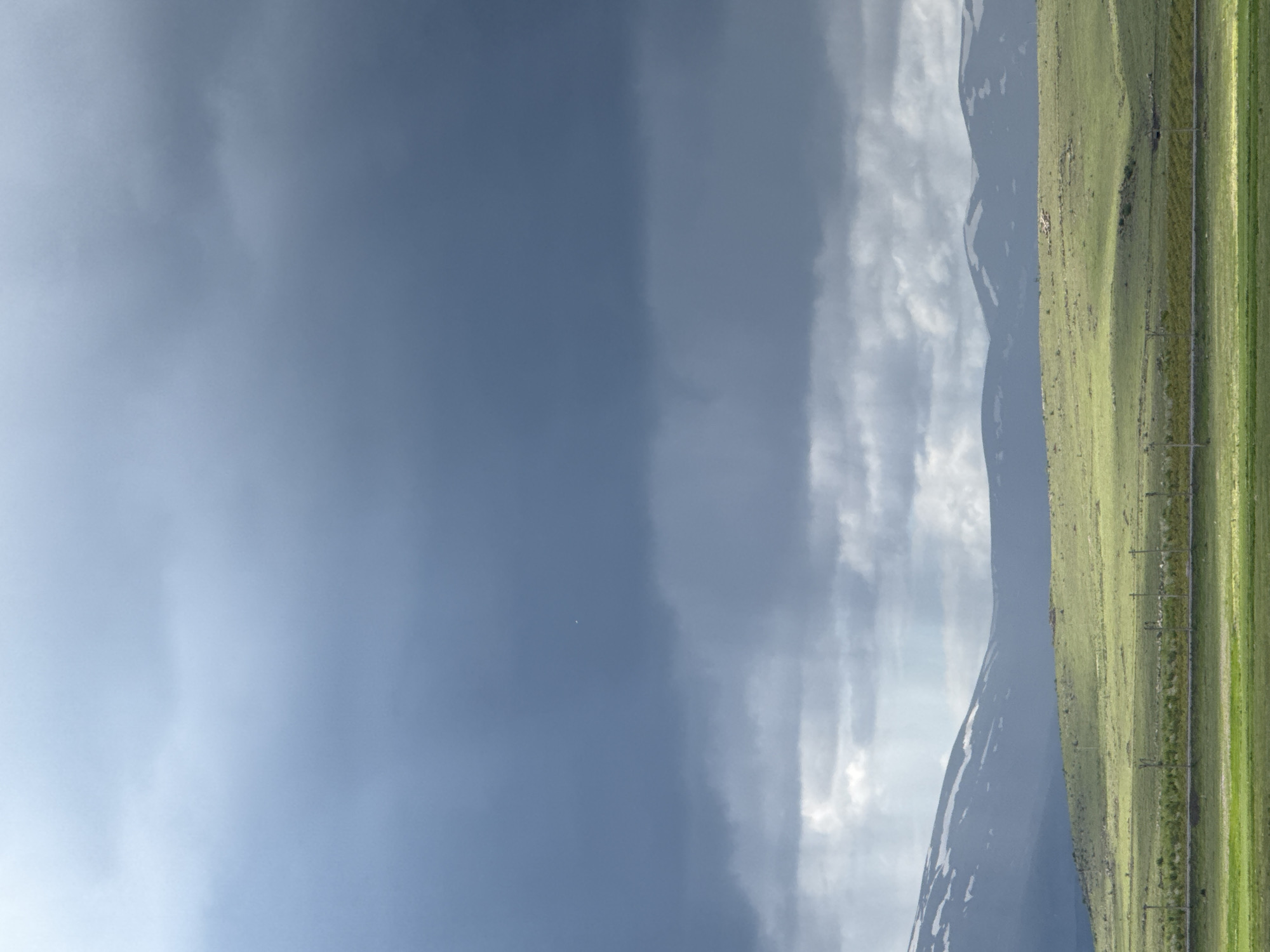 The image depicts a landscape near Tlik, Armenia. The right side of the frame is dominated by a green, grassy field with utility poles running parallel to the edge. Adjacent to the field are mountains with patches of snow. The sky takes up the majority of the frame, ranging from a dark blue and cloudy region to lighter, cloudier area with apparent sun rays.