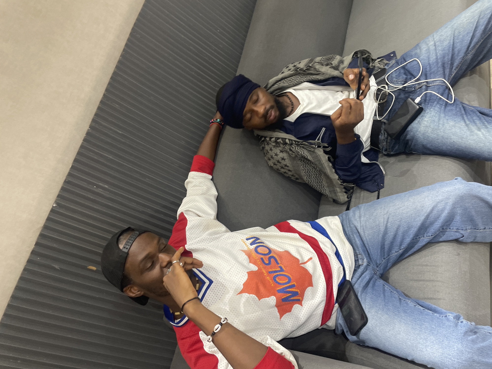Two young men are resting on a gray couch. The man on the left is wearing a black baseball cap, a white and red sports jersey with "MOLSON" and a maple leaf logo, and blue jeans. He has his eyes closed and is touching his face with his right hand, which has rings on two fingers. He is also wearing a black beaded necklace and a bracelet. The man on the right is wearing a dark blue beanie, a white t-shirt, a dark blue jacket with white accents, a grey and black patterned scarf, and blue jeans. He is holding a smartphone in his right hand and a black portable charger is connected to earbuds with a white cable, which are lying on his lap. His eyes are closed, and he appears to be sleeping. The setting appears to be indoors, possibly a living room or lounge area. The couch has a ribbed texture on the side closest to the camera. The overall mood seems relaxed and casual.