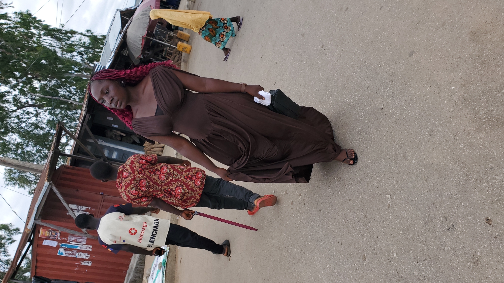 In Jalingo, Nigeria, an outdoor street scene unfolds. A woman with vibrant red braided hair, dressed in a flowing brown gown, walks forward, holding a white object and a black bag. Her gaze is directed to her right. To her left, two men are walking away from the camera. The man on the left wears a white vest with "BALENCIAGA" written on it in black, and black pants. The man to his right is in a red patterned shirt and dark pants, holding a red umbrella. In the background, there are market stalls with tin roofs and various goods, including what appear to be yams. A person in a yellow garment with a patterned skirt is visible walking in the background on the right. The ground is paved with asphalt.