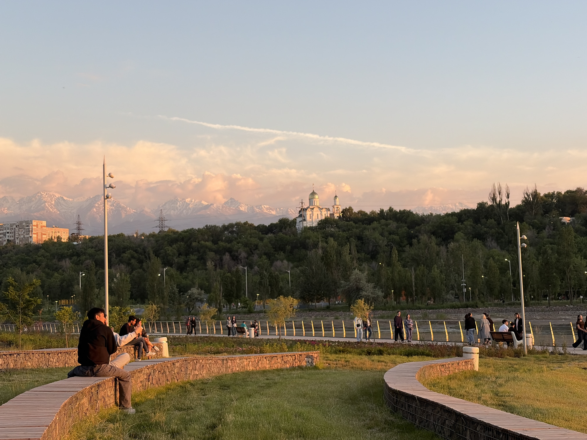 The image shows a public park setting in Almaty, Kazakhstan, at sunset. In the foreground, two young people are sitting on a wooden pathway. One is holding a phone, possibly taking a picture.
In the middle ground, a paved walkway is populated with several groups of people walking and standing. A bench is visible with people seated on it.
In the background, a hill covered with lush green trees rises, and a white Eastern Orthodox church with gold domes sits atop it. Further behind, snow-capped mountains are visible against the sky tinted with warm sunset colors. Electrical towers and a few buildings are also visible in the background.