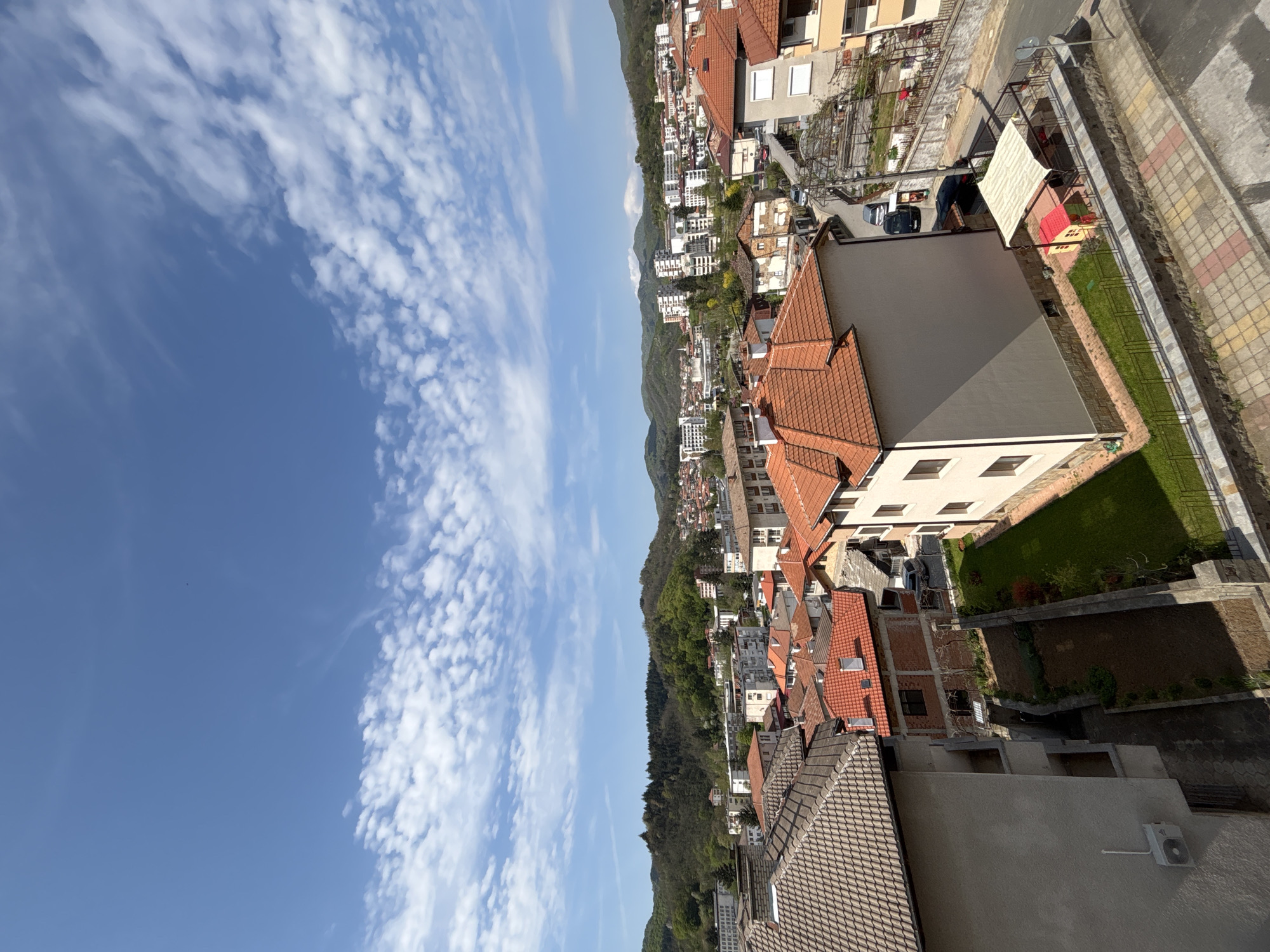An elevated view captures the town of Zlatograd, Bulgaria, sprawling across undulating terrain and integrating with the surrounding green, forested hills. The scene is bathed in bright daylight under a clear blue sky, which is adorned with numerous scattered white cumulus clouds.
The urban landscape is characterized by a dense arrangement of buildings, primarily residential, many featuring distinct red or orange terracotta tile roofs, characteristic of the region. Other buildings exhibit flat grey roofs. The town's structures appear to follow the natural contours of the hills, leading to varied elevations and visible retaining walls. Roads and pathways weave between the buildings, with some cars parked along the streets.
In the foreground, details include a paved area with a colorful, geometric pattern of yellow, red, grey, and white tiles. Adjacent to this, a small grassy patch hosts a white canopy structure and a colorful red and yellow children's playhouse, suggesting a private garden or play area. An outdoor air conditioning unit is visible on the wall of a building. The lush, vibrant green of the forests on the hills forms a natural backdrop, contrasting with the man-made structures.
No specific activities or events are taking place, and no people are clearly visible. There is no discernible text within the image. The time of day appears to be sunny midday or early afternoon, indicated by the bright, clear light and visible shadows.