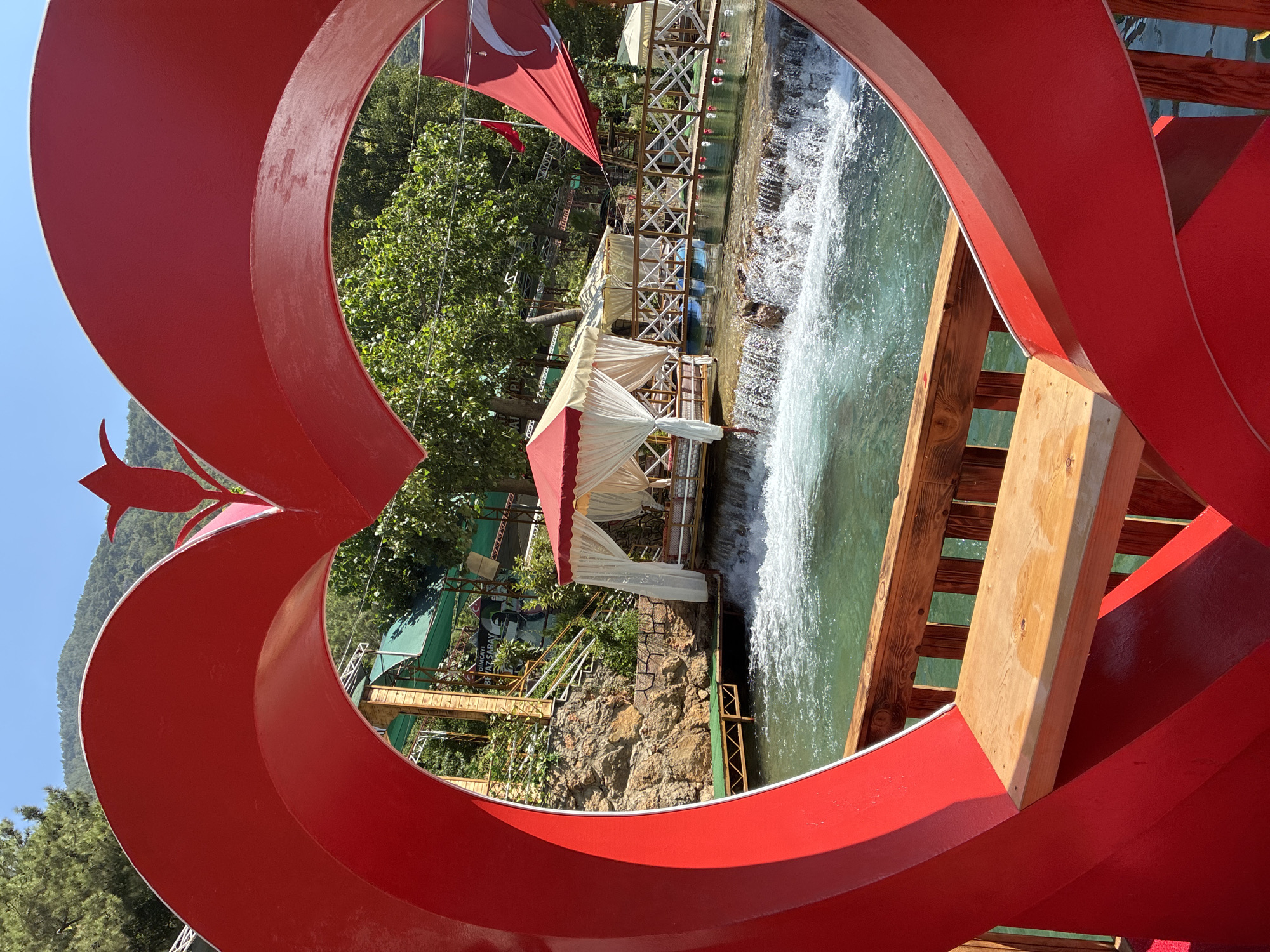 An outdoor scene in Alanya, Turkey, is depicted through a large, red, heart-shaped frame, complete with a wooden bench. The foreground is dominated by the red heart structure, with a small portion of a wooden railing visible. Beyond the heart, a clear, turquoise river flows over rocks, creating a small waterfall. Several gazebos with white curtains and red canopies are situated along the riverbank, some appearing to be seating areas. A wooden bridge with white railings spans the river in the mid-ground. Lush green trees and a rocky hillside form the backdrop, with a clear blue sky overhead. The Turkish flag is visible, flying from a pole near one of the gazebos. The overall ambiance suggests a scenic and tranquil location, possibly a park or a riverside restaurant.