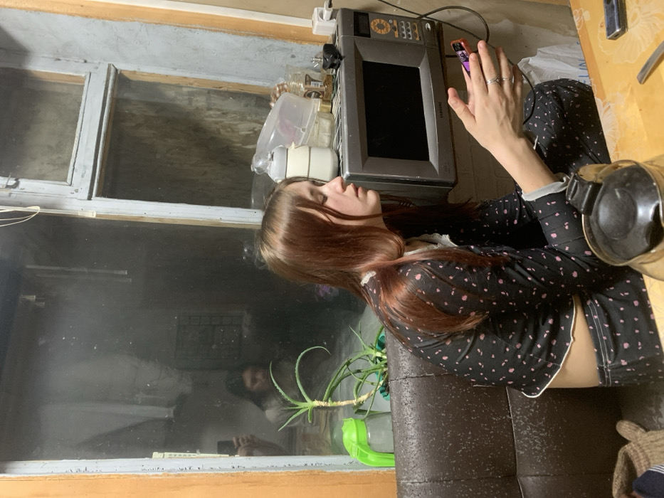 A young woman with auburn hair sits in a chair by a window. She's wearing a black top with pink polka dots. Beside her on a table is a kettle. A microwave sits nearby. Through the window's reflection, another person can be seen holding a phone. A potted plant rests on the windowsill. The scene seems to be set in a kitchen or living area.
