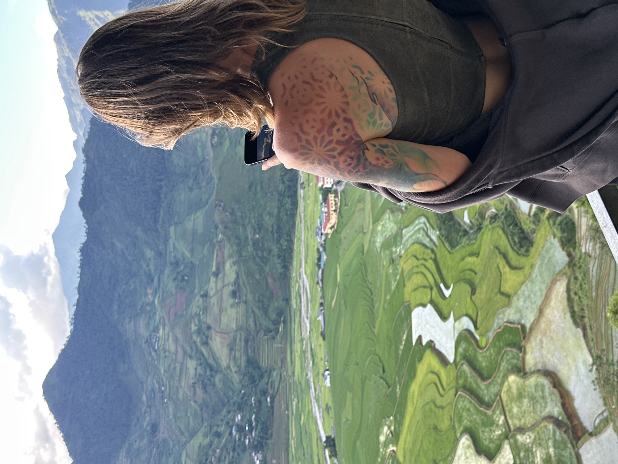 An elevated view captures a person from behind, looking out over the terraced rice fields of Tú Lệ, Vietnam. The individual has medium-length brown hair and is wearing an olive green tank top, with a dark grey zippered hoodie draped over their shoulders. A prominent, multi-colored tattoo featuring geometric and organic patterns is visible on their right shoulder and upper arm. The person holds a black smartphone in their right hand, its screen reflecting the overcast sky. Below, lush green rice paddies, some filled with water reflecting the sky, cascade down the valley, following the contours of the land. A light-colored river or stream winds through the valley floor, with scattered buildings and small villages nestled among the fields. In the distance, multiple layers of verdant, forested mountains rise under a cloudy sky, with hints of blue sky overhead. A metal railing or ledge is partially visible in the lower right foreground, indicating an observation point.