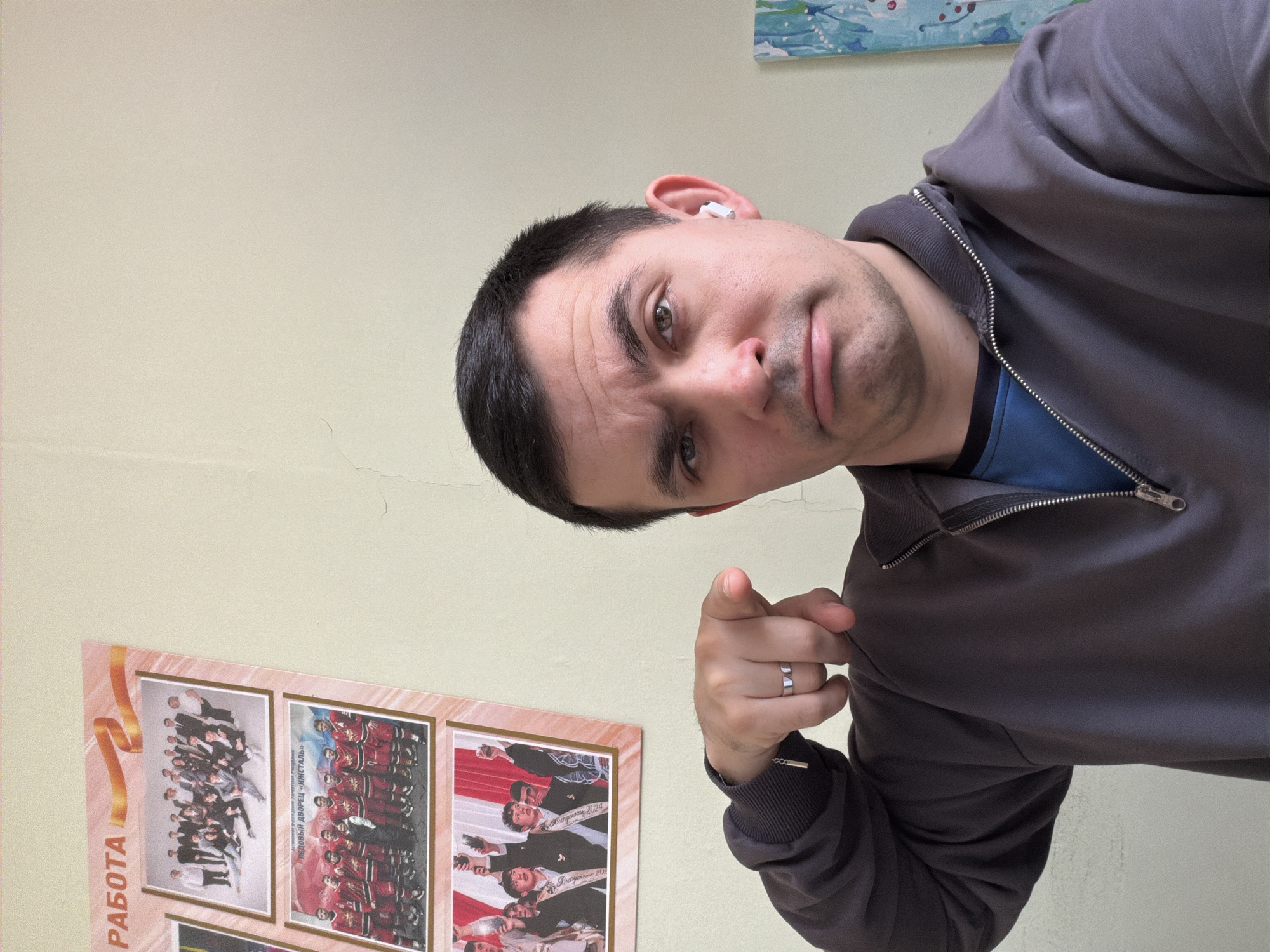 A young man with short dark hair, dark eyes, and light stubble is captured in a medium close-up, leaning into the frame. He wears a dark grey zip-up jacket over a blue shirt, and a white wireless earbud is visible in his right ear. A silver-colored ring adorns the ring finger of his left hand, which is extended and pointing directly forward with the index finger. His expression is direct and focused, with a slight furrow in his brow and a serious gaze towards the viewer.
The background consists of a light cream-colored wall, showing signs of minor cracks in places. On the lower left, several framed images or posters are visible, featuring groups of people. Prominent Russian text can be deciphered on these posters, including "РАБОТА" (Work), "ЛЕДОВЫЙ ДВОРЕЦ «ИЖСТАЛЬ»" (Ice Palace "Izhstal"), and "Выпускники 2024" (Graduates 2024). Part of a blue and white painting or artwork is visible on the upper right side of the wall. The indoor setting, along with the specific Russian place names, aligns with the context of Izhevsk, Russia. The images suggest a public or institutional environment, possibly a school, sports facility, or workplace. There are no direct cues for the time of day or weather.