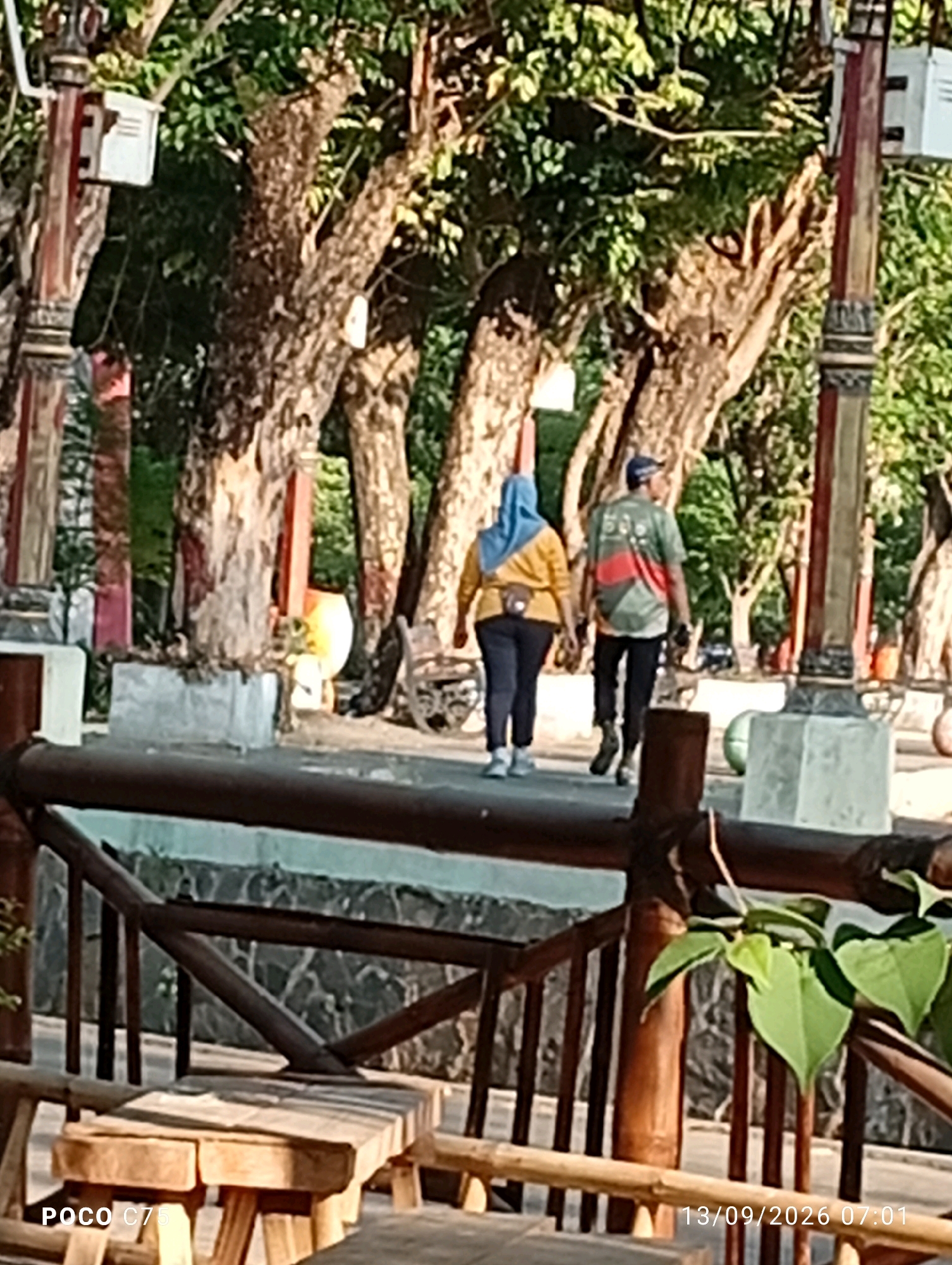 In Jepara, Indonesia, two individuals walk away from the camera in a park-like setting. The person on the left wears a blue hijab and a yellow long-sleeved shirt with dark pants. The person on the right, wearing a dark cap and a green shirt with red and white accents, walks slightly ahead. The environment features large trees with textured bark, a stone wall, and a wooden railing. The lighting suggests it is daytime, with dappled sunlight filtering through the leaves. A wooden bench is visible in the foreground.