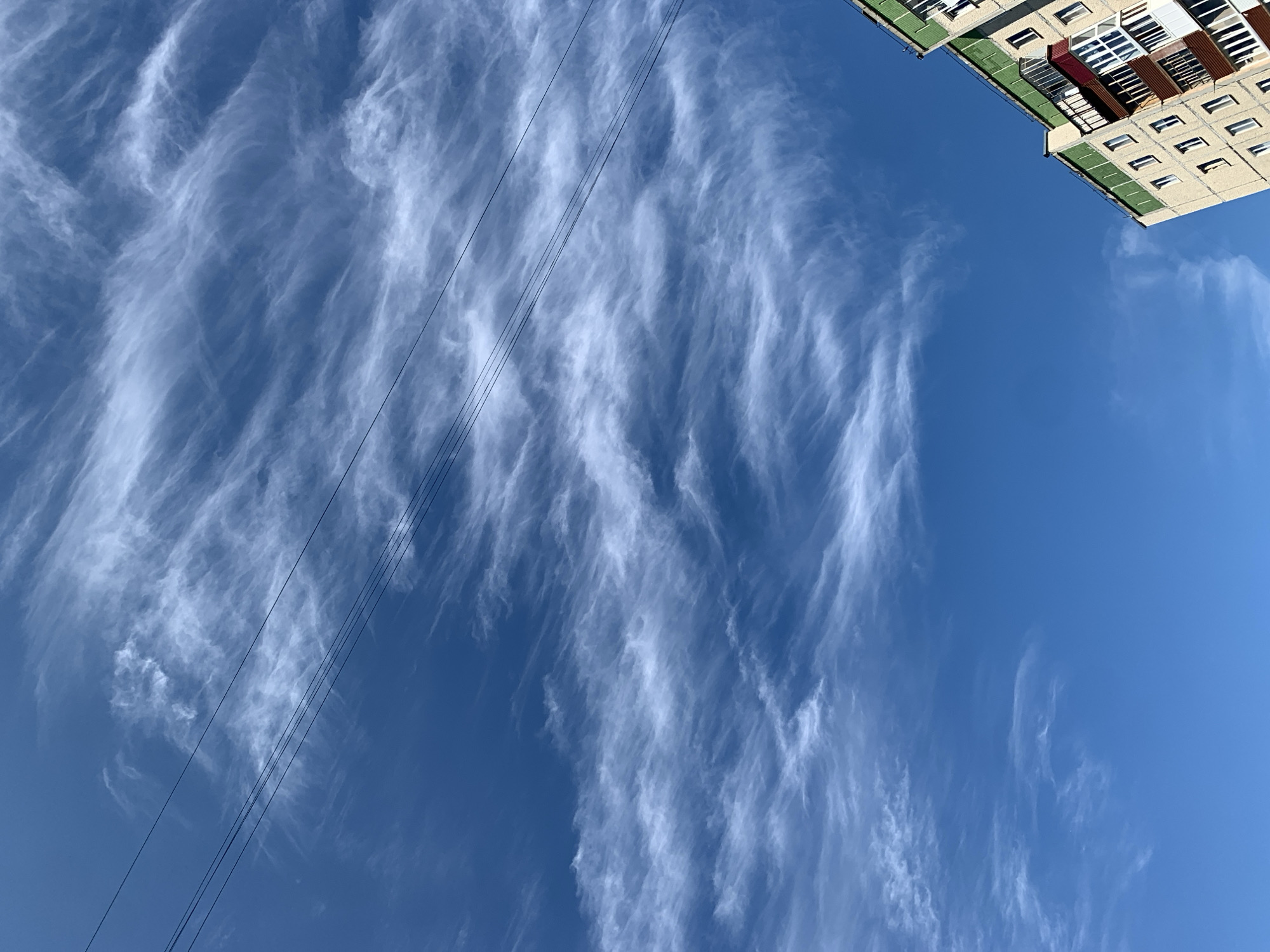 A low-angle shot shows the sky above a multi-story apartment building in Asha, Russia. The sky is a clear, deep blue, with thin, wispy cirrus clouds stretching across it. Several parallel black power lines are visible, running horizontally across the upper half of the frame. The building, with its light-colored facade and green trim, occupies the lower right portion of the image. Its windows are visible, as are several balconies, some enclosed and others open. The building's perspective suggests it is viewed from below and to the side, emphasizing the vastness of the sky. The scene is devoid of people and any immediate action, creating a still and observational mood.