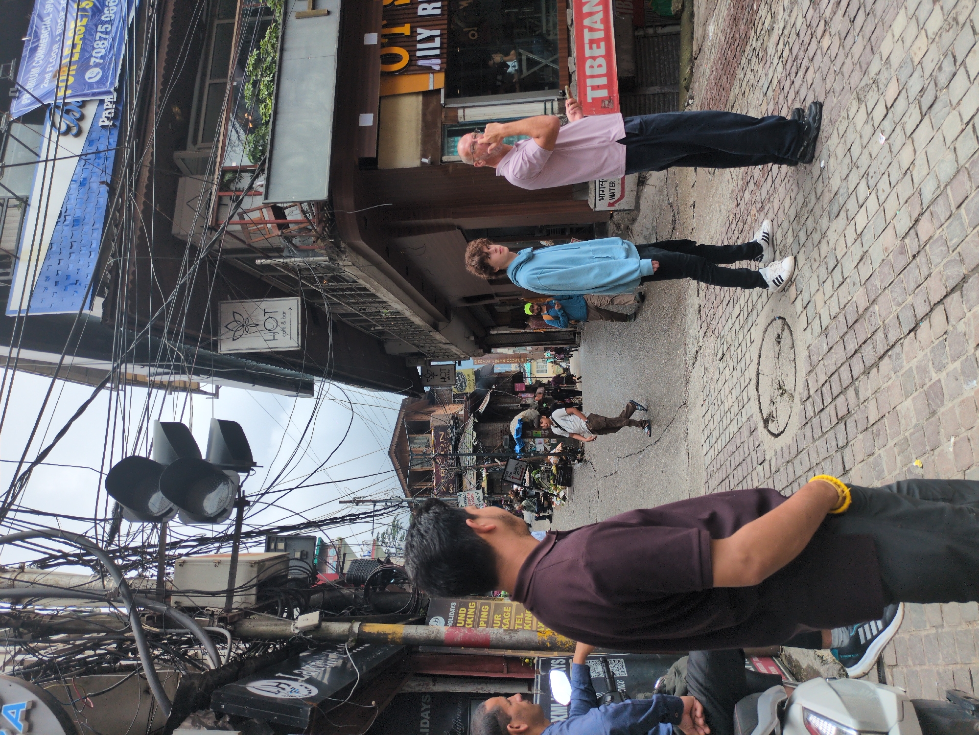 The image captures a street scene in Bhagsu, India, characterized by a dense network of electrical wires overhead and a cobblestone street. In the foreground, a man wearing a maroon polo shirt and black pants stands with his right hand in his pocket, a yellow bracelet visible on his wrist. To his left, another man in a blue shirt is partially visible. Further down the street, a young person in a light blue hoodie and black pants walks away from the camera, while an older man in a pink polo shirt and dark pants stands facing left, his hand near his face. Other individuals are dispersed throughout the scene, including a child in a white shirt and brown pants walking towards the viewer. Buildings line both sides of the street, with various signs and shopfronts visible. A traffic light hangs precariously from a pole amidst the tangled wires. The overall atmosphere appears to be a busy, somewhat chaotic urban environment.