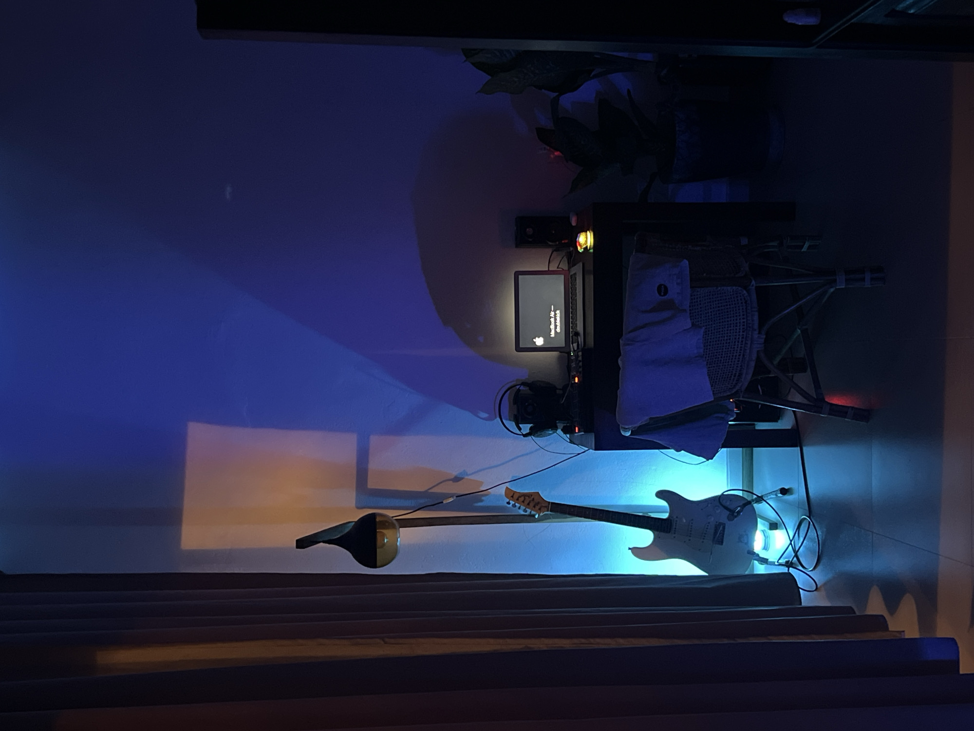 The image shows a desk setup in a dimly lit room, likely an indoor space in Goviyapana, Sri Lanka, based on the prompt. The setup includes a laptop displaying a startup screen, headphones, an electric guitar, and what appears to be a sitar or similar stringed instrument resting on the floor. The desk is against a white wall, illuminated by blue and yellow light, creating shadows. Curtains or blinds are visible in the foreground. There are no people visible in the image. A plant sits on the desk.