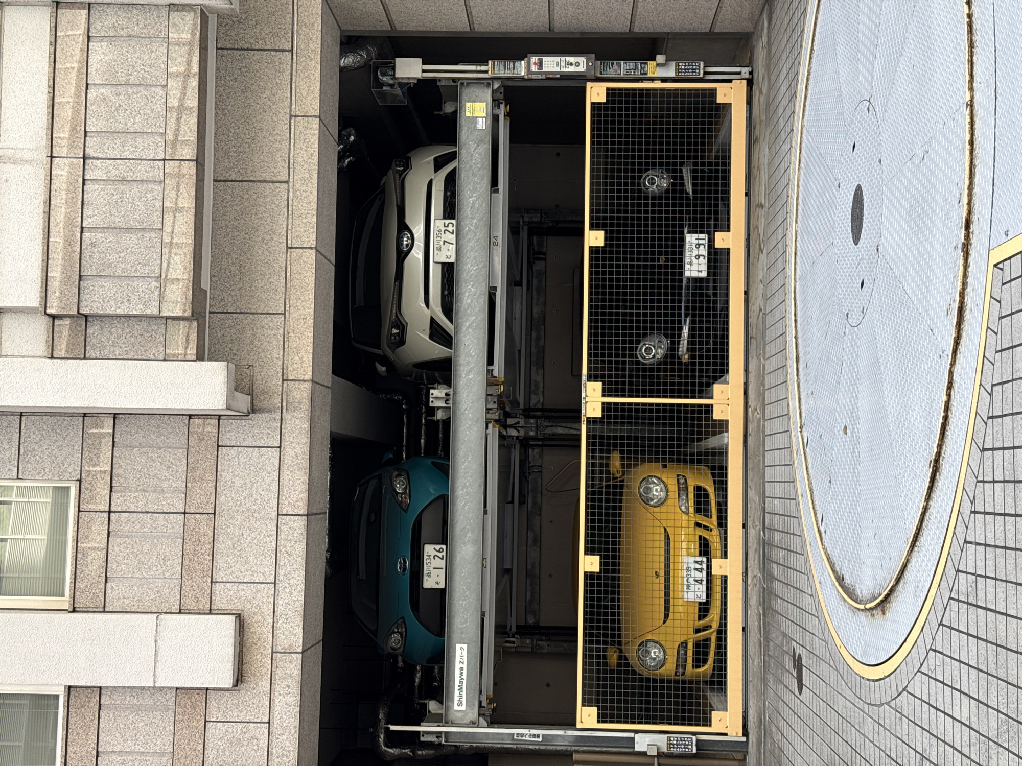The image presents an aerial view of a mechanical parking system in what appears to be an urban environment, likely in the city of Shibuya, Japan. The system houses four cars of various colors: white, teal, black, and yellow, each enclosed within a grid-like frame. A central vertical pillar seems to support the structure. Adjacent to the parking structure is a circular platform, possibly a turntable or rotating stage. The surrounding architecture is predominantly composed of tiled facades, indicating a building structure. License plates are visible on each vehicle, written in Japanese.