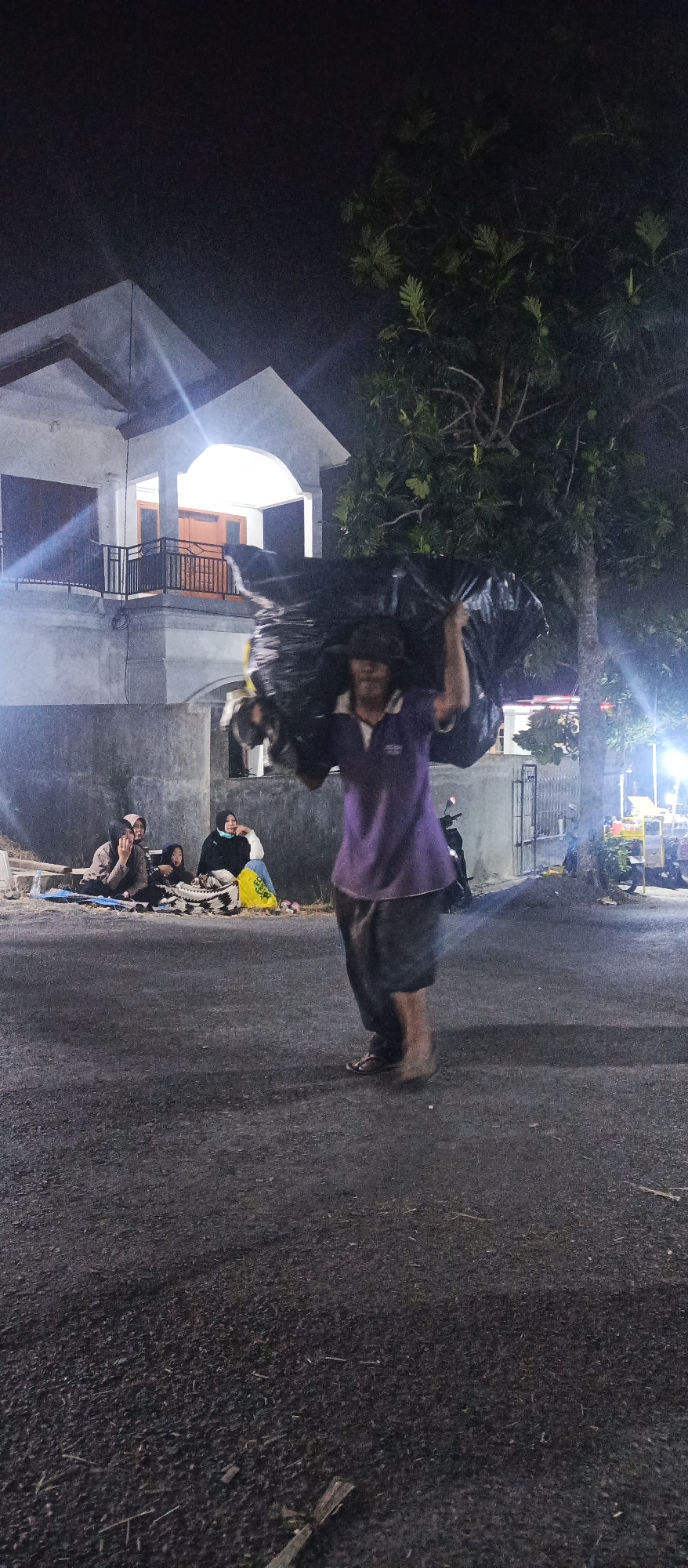 In Sumbersekar, Indonesia, at night, a person wearing a purple shirt and dark pants carries a large black plastic bag over their shoulders. In the background, several people are seated on the ground, some on blankets. A two-story building with a light-colored facade and dark trim is visible behind them, featuring an arched entryway and a balcony. A large tree with dark green foliage stands to the right of the building, and a fence is also present. The scene appears to be on a paved road or area.