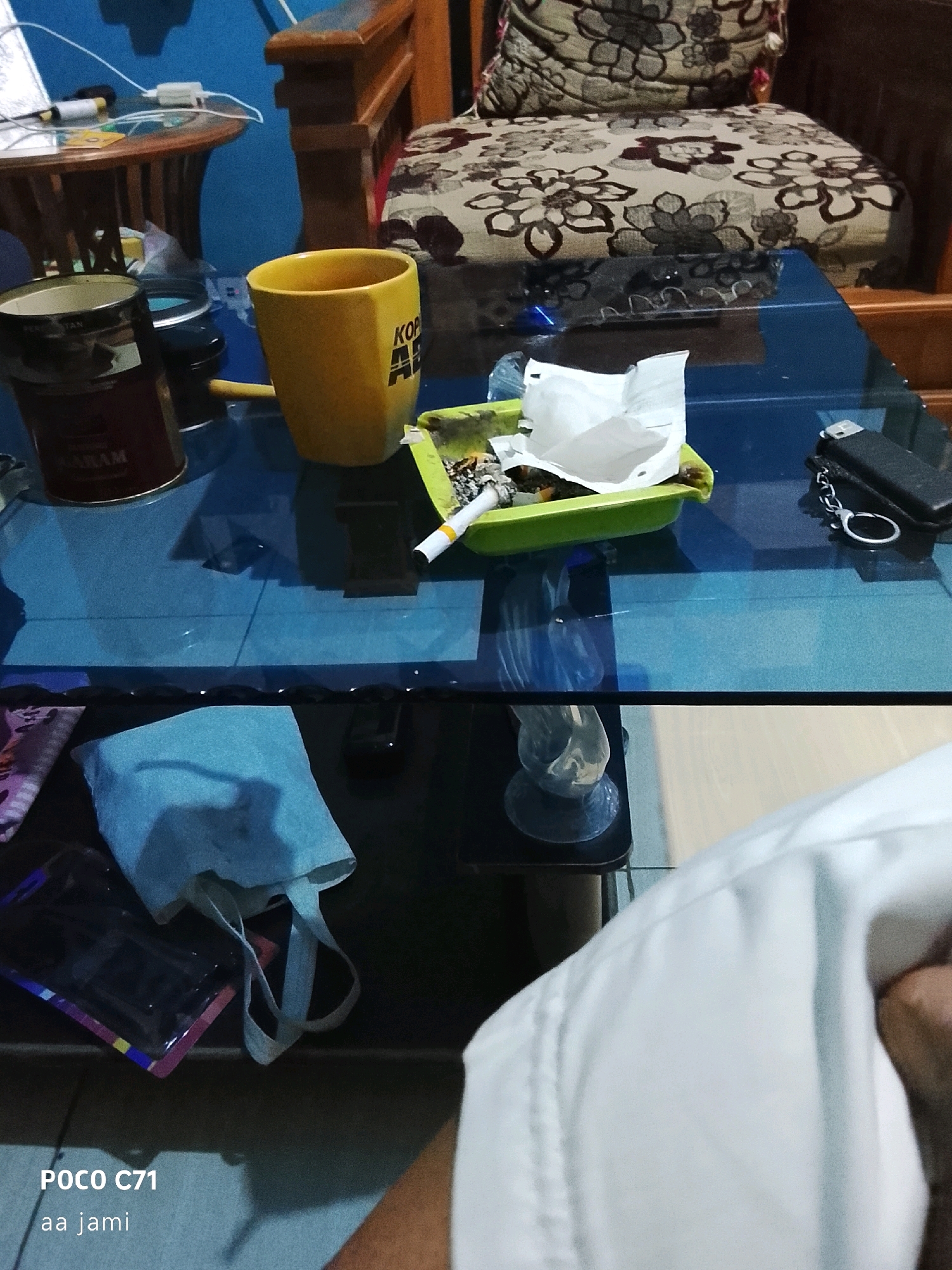 The image shows a close-up of a glass-top table in what appears to be a residential setting in Tasikmalaya, Indonesia. The table is cluttered with various items including a yellow mug with the word "KOP" visible, a metal tin with "MARAM" written on it, a green ashtray containing cigarette butts and a crumpled piece of white paper, a lighter with a keychain, a cell phone, and a light blue cloth bag with handles. In the background, a wooden chair with a floral patterned cushion is visible, as well as a smaller round wooden table. The lighting is dim, suggesting it may be indoors or late in the day. The lower portion of the image shows a person's white shirt sleeve and hand. The text "POCO C71 aa jami" is overlaid on the bottom left corner, indicating the device used to capture the image and possibly a username.