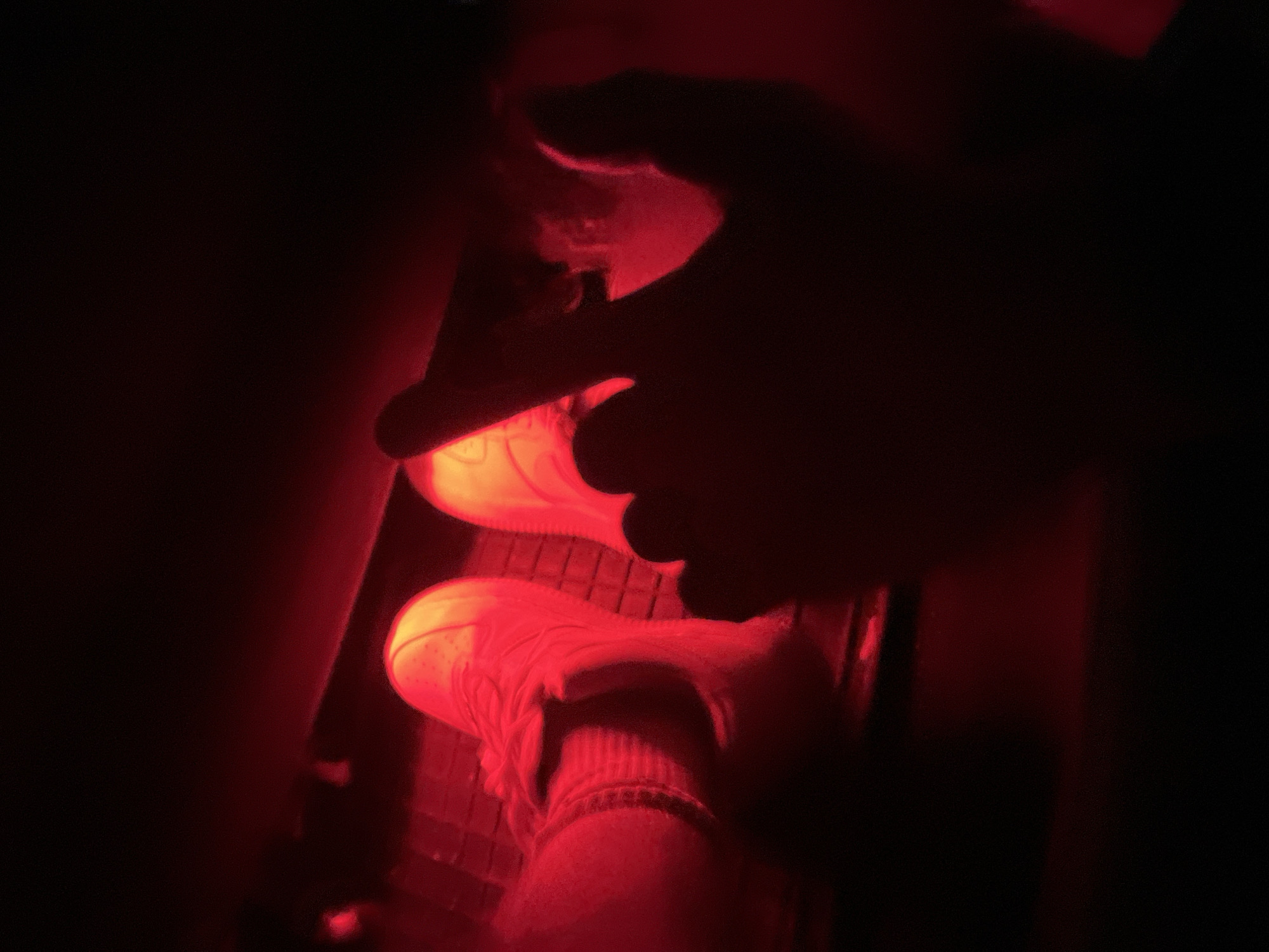 In a dimly lit environment, illuminated by a red light source, a pair of feet wearing white sneakers with ribbed socks are visible. The sneakers appear to be resting on a textured surface, possibly pedals. A human hand, also silhouetted and illuminated by the red light, is positioned in the foreground, with its index finger extended towards the shoes. The background is predominantly dark, with hints of red light reflecting on surfaces. The scene suggests a clandestine or atmospheric setting, possibly within a vehicle given the presence of pedals. The location is not identifiable from the image alone; however, considering the request for Zlatograd, Bulgaria, this scene could be interpreted as occurring within a private residence, a vehicle, or a dimly lit entertainment venue in that city.