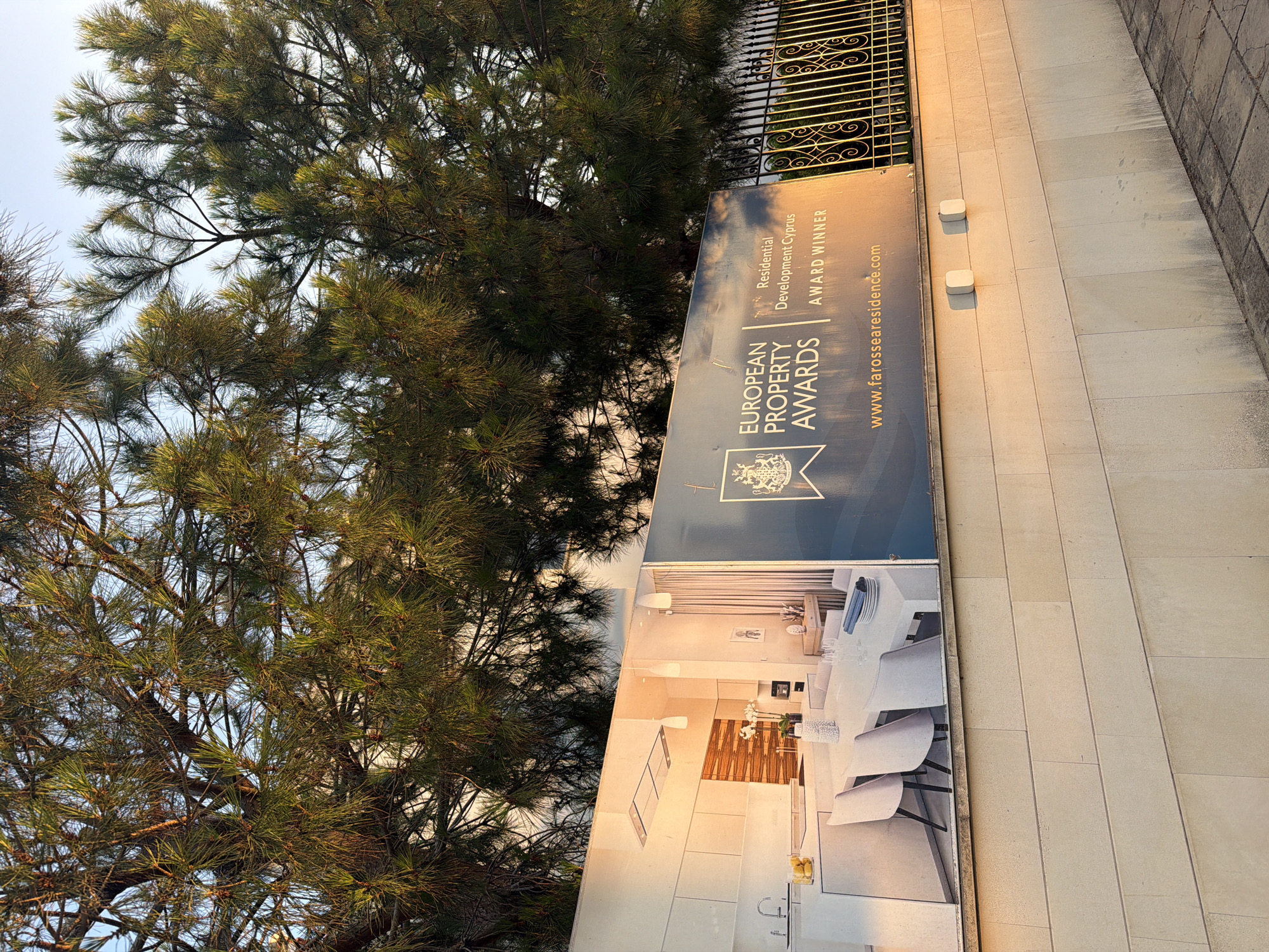 The image captures a billboard advertising a residential development in Pervolia, Cyprus, set against a backdrop of lush green pine trees and a bright, clear sky. The billboard prominently displays "EUROPEAN PROPERTY AWARDS RESIDENTIAL DEVELOPMENT CYPRUS AWARD WINNER" alongside a pristine image of a modern kitchen and dining area. The text "www.farossea.com" is also visible at the bottom of the billboard. To the right, a decorative wrought-iron fence adds a touch of elegance to the scene. The billboard is mounted on a light-colored stone wall, which is illuminated by the warm glow of the setting sun, casting soft shadows. The foreground shows a paved walkway with some dirt and stains, suggesting an outdoor urban environment. The overall impression is one of a sophisticated and successful real estate project being showcased in a pleasant, sunny location.