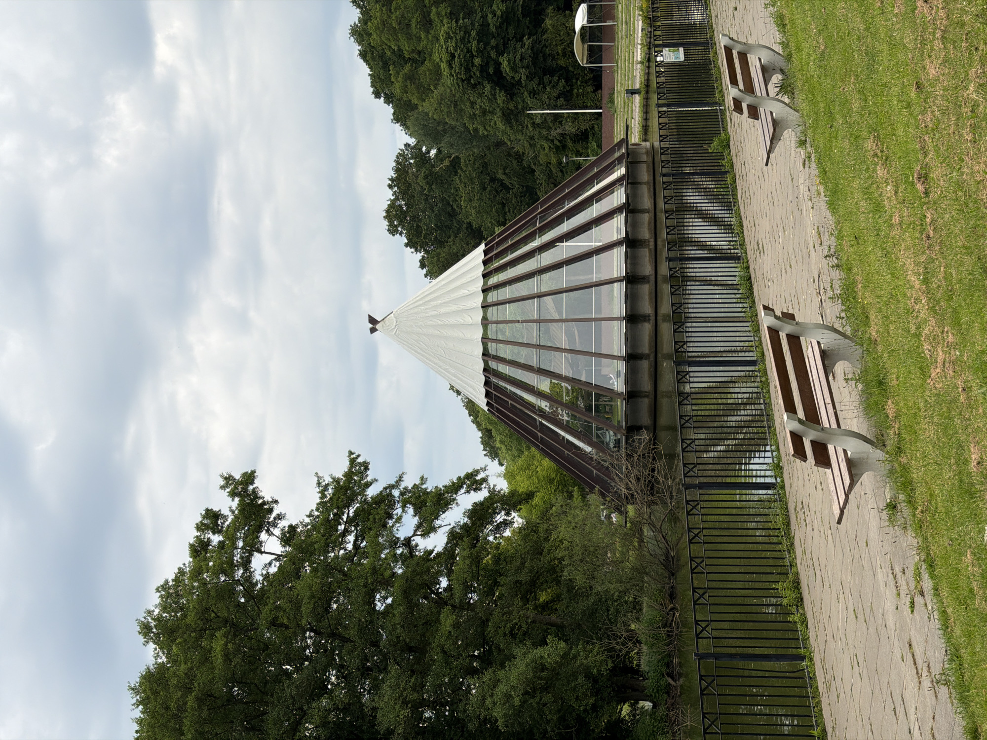 The image depicts a conical structure with a glass base and a white tarp roof, situated on a body of water. The structure is surrounded by lush green trees. In the foreground, a paved walkway runs alongside the water, bordered by a black metal fence. Two park benches are placed on the walkway, facing the water and the conical structure. The sky is overcast with gray clouds. The scene appears to be a park or recreational area, possibly in the city of Helmond, Netherlands, given the architectural style and landscape.
