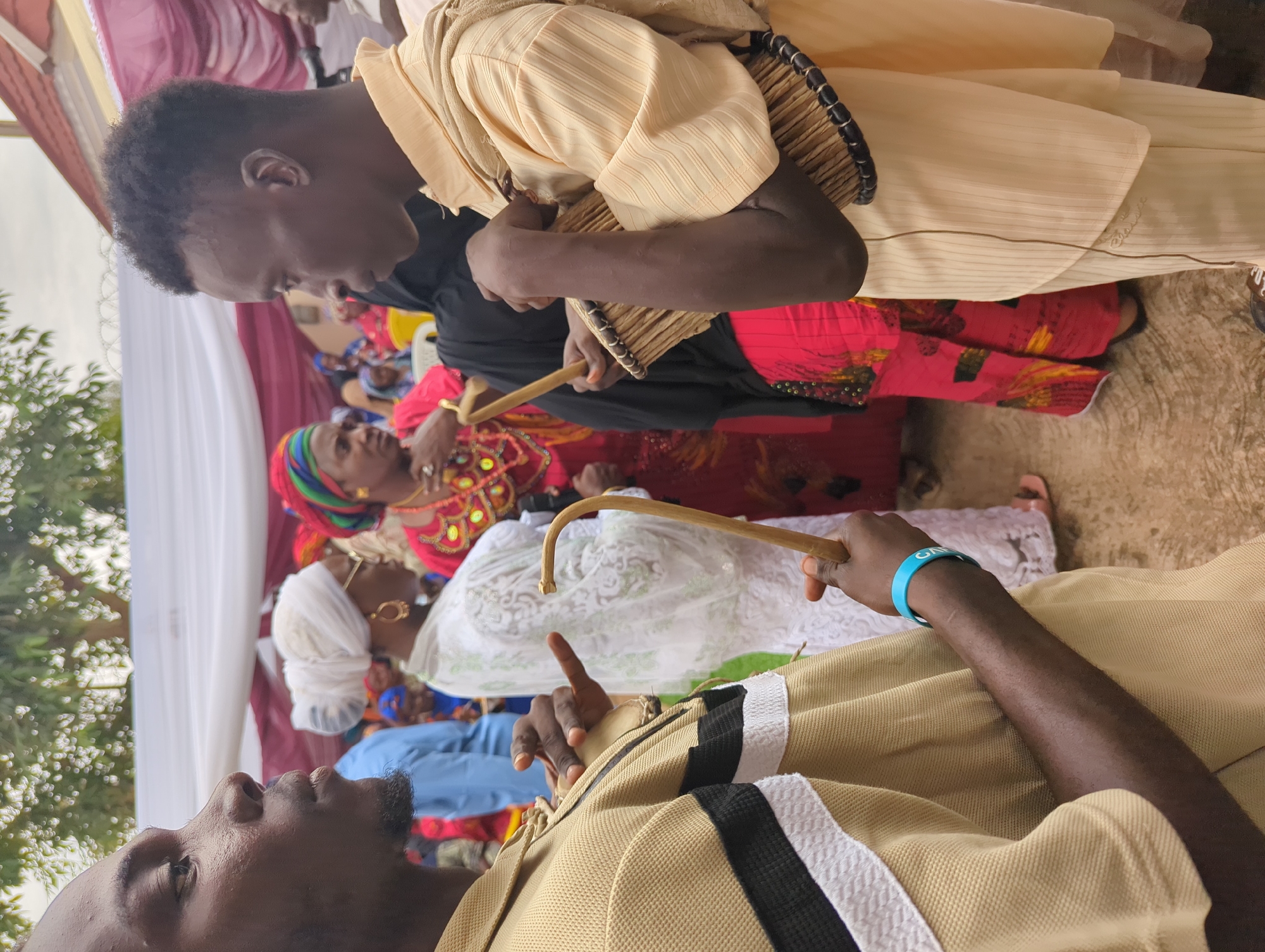 This image depicts an outdoor gathering in Unguwar Rimi, Nigeria.
In the left foreground, an adult male with a dark complexion is seen in profile, facing right. He has short dark hair and a light beard. He wears a tan polo shirt with a horizontal black and white stripe across the chest. His left hand holds a small, light-colored hand drum, and his right hand holds a curved wooden beater. A blue wristband is on his right wrist.
In the right foreground, a second adult male with a dark complexion is also in profile, facing left, towards the first man. He wears a light yellow, vertically striped long-sleeved shirt with a matching fabric draped over his left shoulder and light-colored trousers. He holds a small, light-colored hand drum against his body with his right arm and hand, and a curved wooden beater in his left hand. He has short dark hair.
In the midground, between the two men, an adult female with a lighter complexion is visible in profile, facing right. She wears a white headwrap, a white lace dress, gold hoop earrings, and eyeglasses. To her right, another adult female wears a red headwrap and a red dress with patterned details. The background contains additional individuals, predominantly women in various colorful traditional garments and headwraps, some standing and some seated. A yellow plastic chair is partially visible behind the woman in red.
The background features white and maroon fabric drapes, indicating a temporary canopy or tent structure. Above the drapes on the right, barbed wire fencing is visible. Green tree foliage is present at the top of the frame. The ground is a light-colored, textured surface. The scene is illuminated by diffused natural light, consistent with daytime.