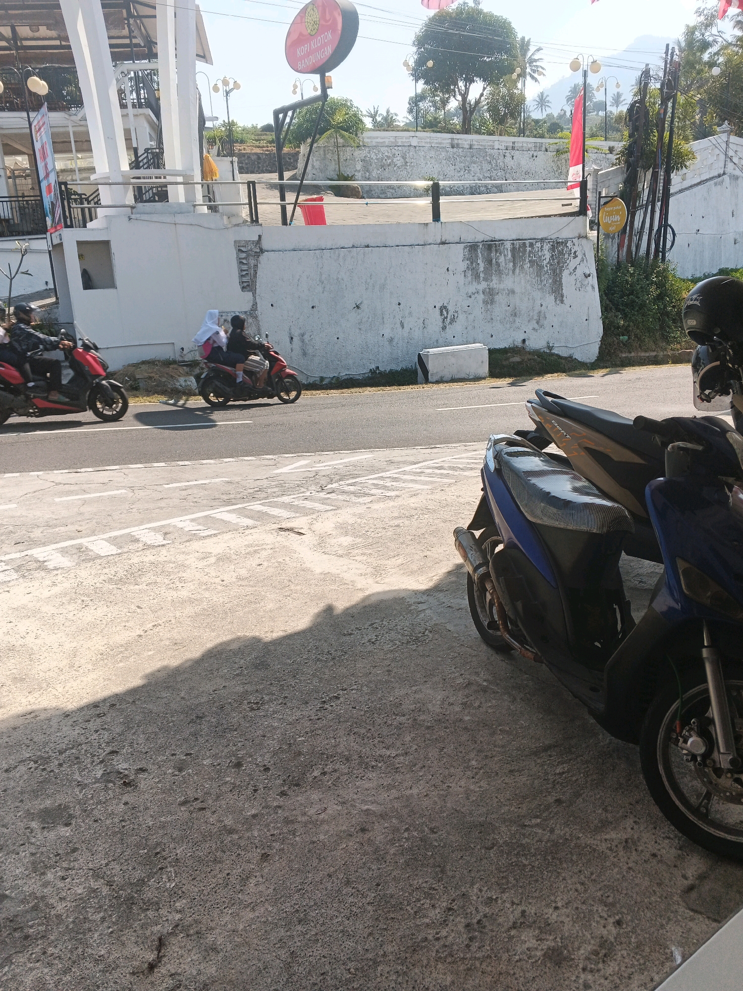 In Pakopen, Indonesia, two motorcycles are seen on the side of a road. The first motorcycle, a red scooter, is ridden by a person wearing a black jacket and helmet, with a white and pink bag on the back. The second motorcycle, also a red scooter, carries two passengers: a person in a white hijab and a person in a black jacket. They are on a road with white lane markings and a shadow cast by a nearby structure. In the background, there is a white concrete wall, a building with white pillars, and trees. A circular red sign with text is visible on a pole. Further in the background, a distant mountain is visible. In the foreground, another motorcycle, a blue scooter, is parked on a textured surface.