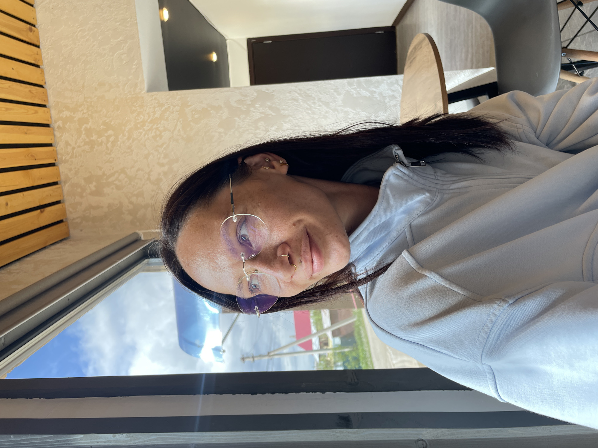 An indoor scene in Oktyabrsky, Russia, features a female subject seated at a window, gazing outdoors. The woman, appearing to be in her 20s or 30s, has dark hair and visible freckles on her face. She wears rimless eyeglasses with a gold-toned frame and subtly tinted lenses. Her facial piercings include a gold hoop in her right nostril and a small labret stud below her lower lip. Her right ear is adorned with multiple small gold-toned earrings. She is dressed in a light-colored, possibly light blue or off-white, zippered jacket with visible seam details.
The indoor environment suggests a cafe or similar establishment. To the woman's right, a textured, light beige wall is visible, transitioning to a dark brown door within a white frame. The ceiling above the textured wall features decorative wooden slats separated by dark strips. A section of the ceiling further back is dark with recessed lighting. A round wooden table with a dark base and a gray modern chair are partially visible to the right of the woman.
Through the large window on the left, an outdoor view is discernible under a blue sky with white clouds. Distant structures include a wooden utility pole with wires, a building with a red sloped roof, and another building with light-colored walls. Green vegetation, indicative of grass and trees, is present, suggesting a suburban or semi-rural landscape. A faint blue reflection is present in the upper portion of the window glass.