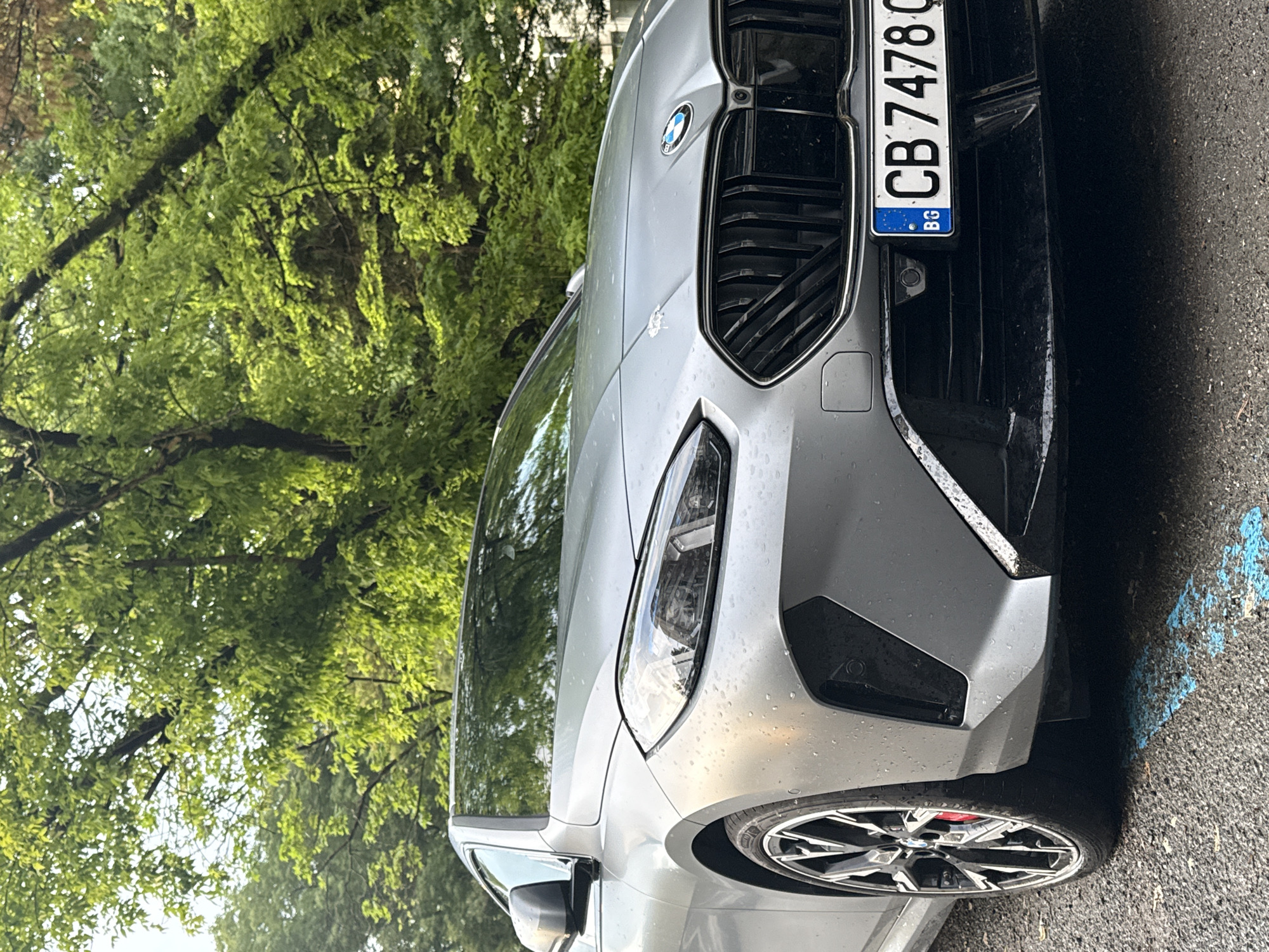 The image shows a gray BMW car parked on an asphalt surface. The car's front grill, headlight, and front tire are visible. The license plate reads "CB 7478 BG." Based on the "BG" country code on the license plate, the location is Bulgaria. Without further information, the specific city of Burgas cannot be confirmed. Green trees are visible in the background. There are blue markings on the asphalt near the car. The car appears to be wet, possibly from rain. No people are visible.