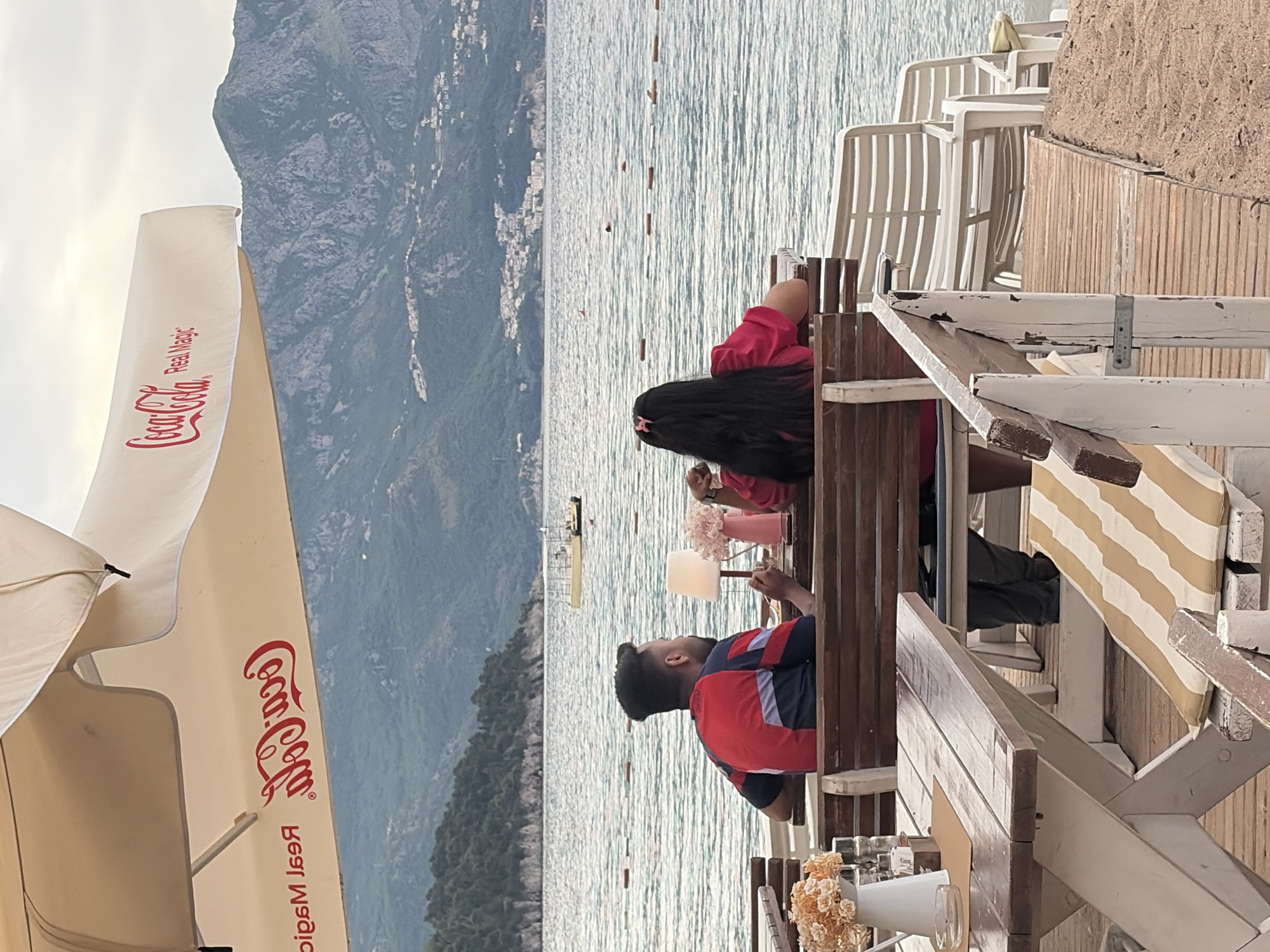 The image captures a scene at a waterfront location in Budva, Montenegro. A man and a woman are seated at a wooden table on a terrace overlooking the Adriatic Sea. The woman, with long, dark hair, is wearing a pink shirt. The man, seen in profile, has dark hair and is wearing a red and blue shirt. They appear to be engaged in a conversation over drinks.
Several tables and white plastic chairs are visible on the terrace. A large white Coca-Cola branded sun umbrella is partially visible on the left side of the image. In the background, the Adriatic Sea stretches out, with mountains rising along the coastline. The mountain range shows clear details of the urban development of the city.