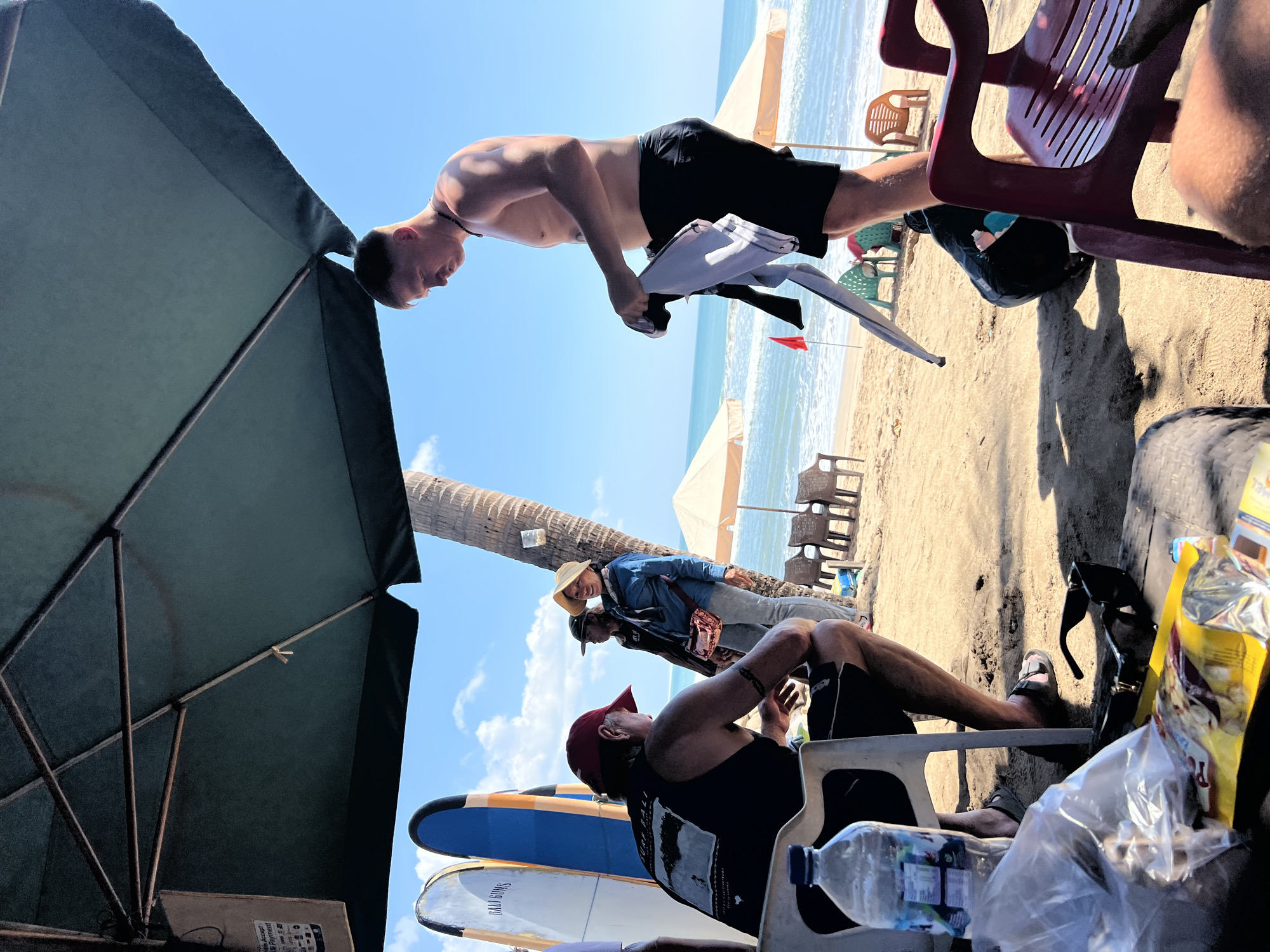 An outdoor scene on the sandy beach of Kuta, Indonesia, is depicted. Several people are present under a large green umbrella, with surfboards leaning against a palm tree. In the foreground, a man sits in a white plastic chair, facing away from the camera, with a bottle of water and snacks on a low table. To the right, a shirtless man in black shorts stands, holding clothing. Further back, near the water's edge, a group of people are gathered, with beach chairs and umbrellas visible. The ocean and sky are clear.