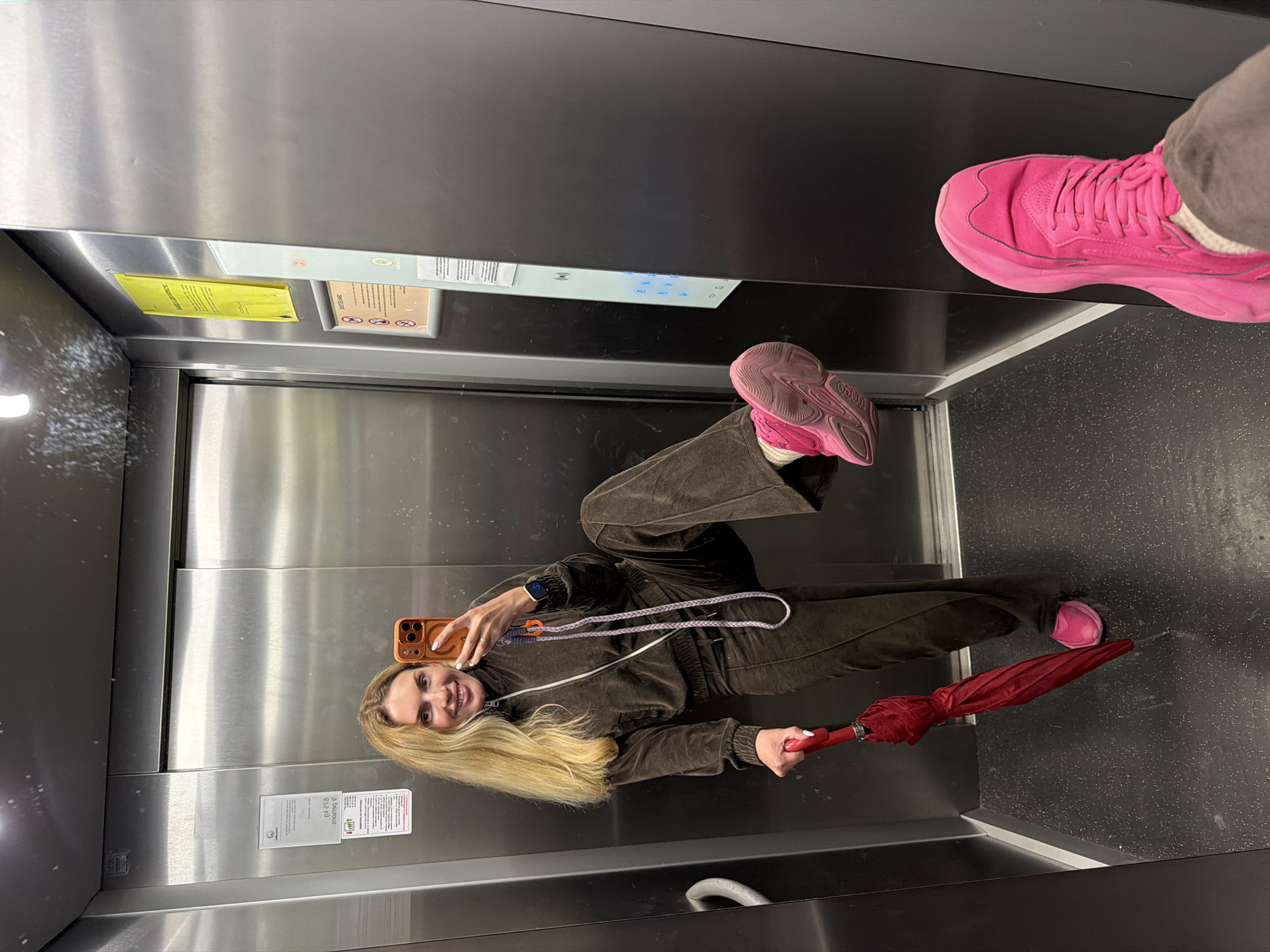 A young woman is taking a selfie in an elevator with stainless steel walls. She is wearing a brown velour tracksuit and pink sneakers, and holding a red umbrella. The elevator has standard signage and buttons, and there is a person's leg with a pink shoe visible at the top of the frame. The exact location in Budva, Montenegro cannot be determined from the image.