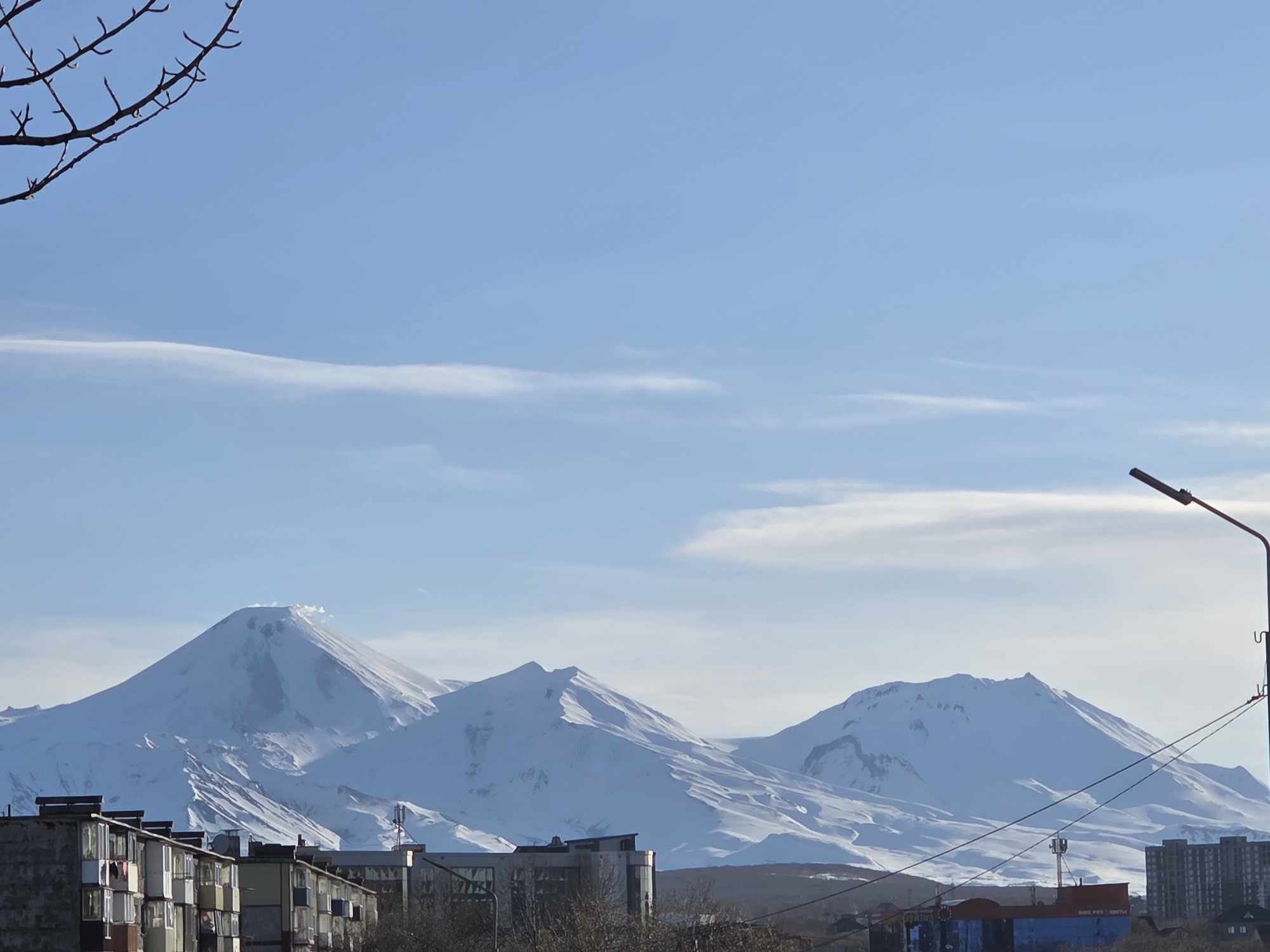 The image depicts a cityscape with snow-capped mountains in the background. The foreground features buildings of various sizes and architectural styles, ranging from older, multi-story apartments with visible wear and tear to more modern structures. A leafless tree branch is visible in the upper left corner, and a street lamp stands on the right side. The sky is blue with some wispy clouds. The mountains in the background are covered in snow, and one appears to be emitting steam or smoke from its peak. The location appears to be within a city, but the specific city is unknown.