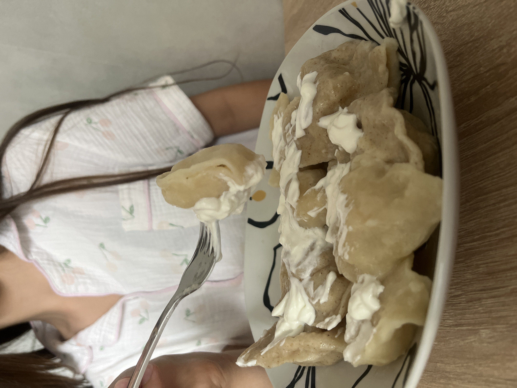 In Rybnitsa, Moldova, a person wearing pajamas with a cherry pattern is eating a plate of dumplings topped with sour cream. The dumplings are light brown and have a slightly wrinkled texture. The plate has a black and white abstract design. The person is holding a fork with one dumpling on it, about to eat it. The background is a plain, light-colored wall and a wooden table.