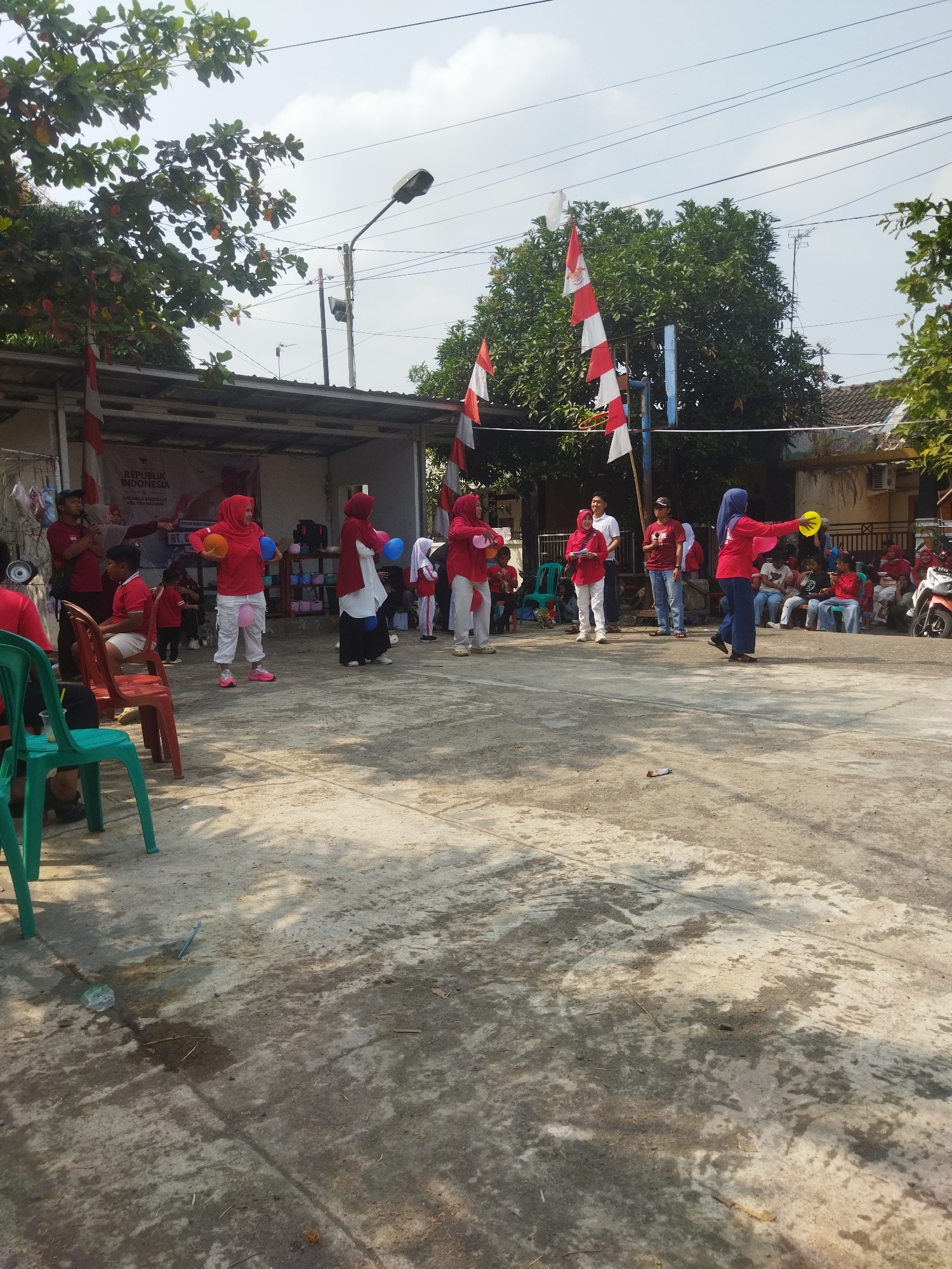 The image depicts an outdoor community gathering, likely a celebration or competition, in Banjar, Indonesia. The scene is set in an open paved area, possibly a courtyard or village square, with a building structure in the background. The building has a corrugated roof and is decorated with red and white Indonesian flags, suggesting a national holiday observance.
Several individuals are participating in an activity involving balloons. Some are holding balloons between their legs, while others appear to be tossing or catching them. A woman in a red hijab and white pants is central to the activity, holding a yellow object in her right hand and a red balloon in her left. Another woman in a blue hijab on the right side of the frame is also holding a yellow object. Many participants and spectators are wearing red, a color commonly associated with Indonesian celebrations.
In the background, there are spectators seated on chairs and benches, some of whom are also dressed in red. A motorcycle is parked on the right side of the image. The ground is concrete and shows signs of wear and tear, with some water stains and debris. Trees with green foliage and some red fruits are visible in the upper left and right portions of the frame, indicating a tropical environment. A streetlamp stands in the background, along with power lines crisscrossing the sky. The sky is mostly clear with some scattered clouds. The overall atmosphere appears festive and communal.
