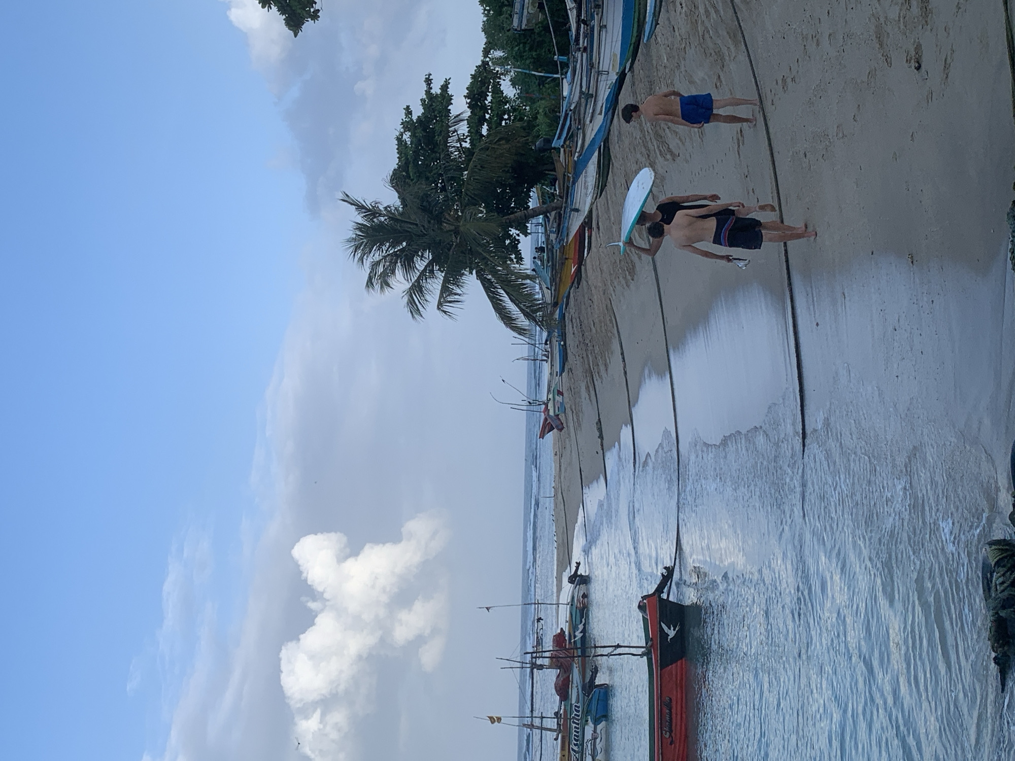 The image depicts a beach scene in Weligama, Sri Lanka. In the foreground, the calm blue ocean laps gently onto a sandy shore. Several colorful fishing boats are moored in the shallow water, some with outboard motors attached. A prominent red and black boat with a white bird emblem is closest to the viewer. The beach itself is lined with more fishing boats pulled up onto the sand.
Further down the beach, three young males are walking away from the water. The one in the middle is carrying a light blue surfboard above his head. He is wearing a black tank top and dark shorts. The male to his left is wearing dark patterned shorts and appears to be holding something in his right hand. The male to his right is wearing blue swim shorts.
In the background, the ocean stretches to the horizon under a partially cloudy sky with patches of blue. A palm tree and other lush green vegetation are visible along the coastline. The overall impression is a peaceful, coastal environment in Sri Lanka, likely at the beginning or end of a day of fishing or surfing.