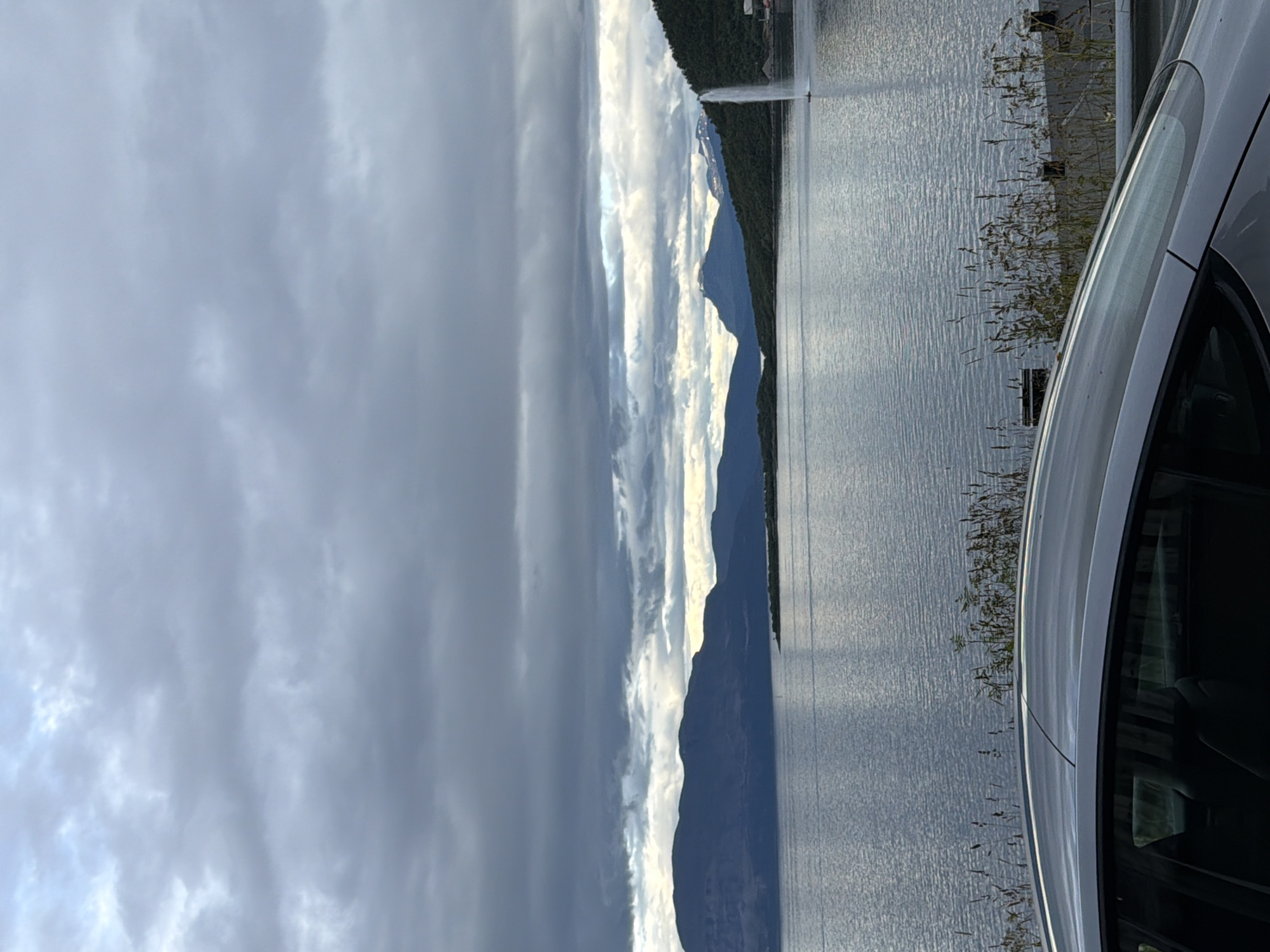 The image depicts a scenic view of a body of water with mountains in the background. A fountain is visible spraying water into the air on the right side of the frame. In the foreground, the roof and side of a silver car are visible. The sky is overcast with dramatic clouds. This view is consistent with locations around Harstad, Norway, specifically the Harstad Marina area, where a large fountain is a prominent feature.