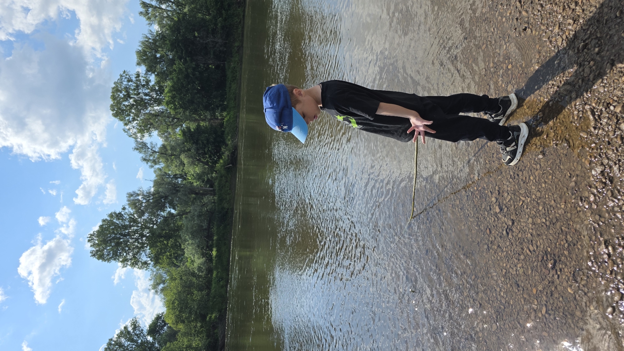 A young boy wearing a blue baseball cap, a black t-shirt with a green graphic, black pants, and sneakers stands at the edge of a shallow river in an outdoor environment. He is holding a thin stick, angled towards the water, and looking down at it. The riverbed is visible through the clear, shallow water, composed of small pebbles and rocks. Lush green trees and foliage line the opposite bank of the river, under a bright blue sky with scattered white clouds. The scene appears to be a natural setting, possibly a park or a natural area, not within a specific urban city location.