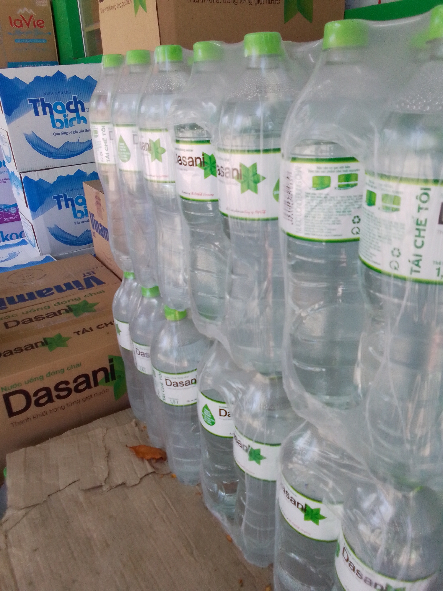 The image shows several rows of bottled water products, mostly Dasani brand, stacked and partially wrapped in plastic. The bottles are clear plastic with green caps and white labels featuring green accents and text in what appears to be Vietnamese. Some bottles are in clear plastic shrink wrap, forming multi-packs. In the background, there are cardboard boxes, one prominently displaying the "LaVie" brand logo and text, and another with "Thạch bích" branding. A brown cardboard box with "Dasani" branding and Vietnamese text is visible on the lower left, showing the text "NƯỚC UỐNG ĐÓNG CHAI" (Bottled Drinking Water) and "Thanh khiết trong từng giọt nước" (Pure in every drop of water). The overall environment suggests a retail or storage area for beverages. The presence of specific Vietnamese brands like LaVie, Thạch bích, and Dasani, commonly sold in Vietnam, along with the Vietnamese language text, indicates the location is likely within Vietnam. Given the prompt's request to include the city of Đà Nẵng, it is plausible this scene is in Đà Nẵng, Vietnam, though the image itself does not contain explicit geographical markers for this specific city.