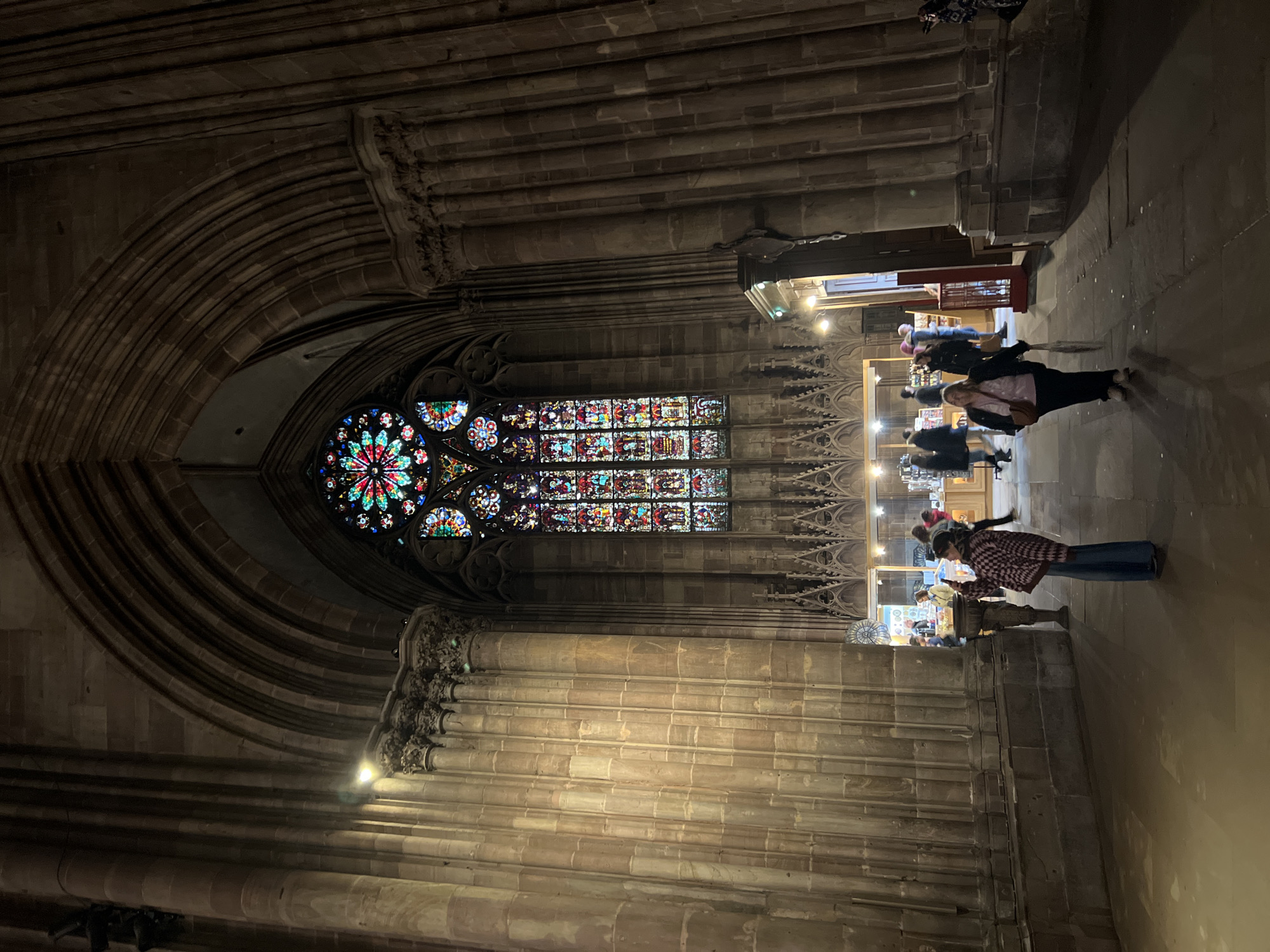 The image captures the interior of the Cathédrale Notre-Dame de Strasbourg in Strasbourg, France, at night. A large, ornate stained-glass window is the focal point, with a circular rose window above a rectangular panel of stained glass. The architecture is Gothic, with ribbed vaults and sculpted stone details. Several people are present on the right side of the frame, some appearing to be browsing in a shop or market area. The overall lighting is dim, with artificial lights illuminating the shop area and highlighting the stained glass.