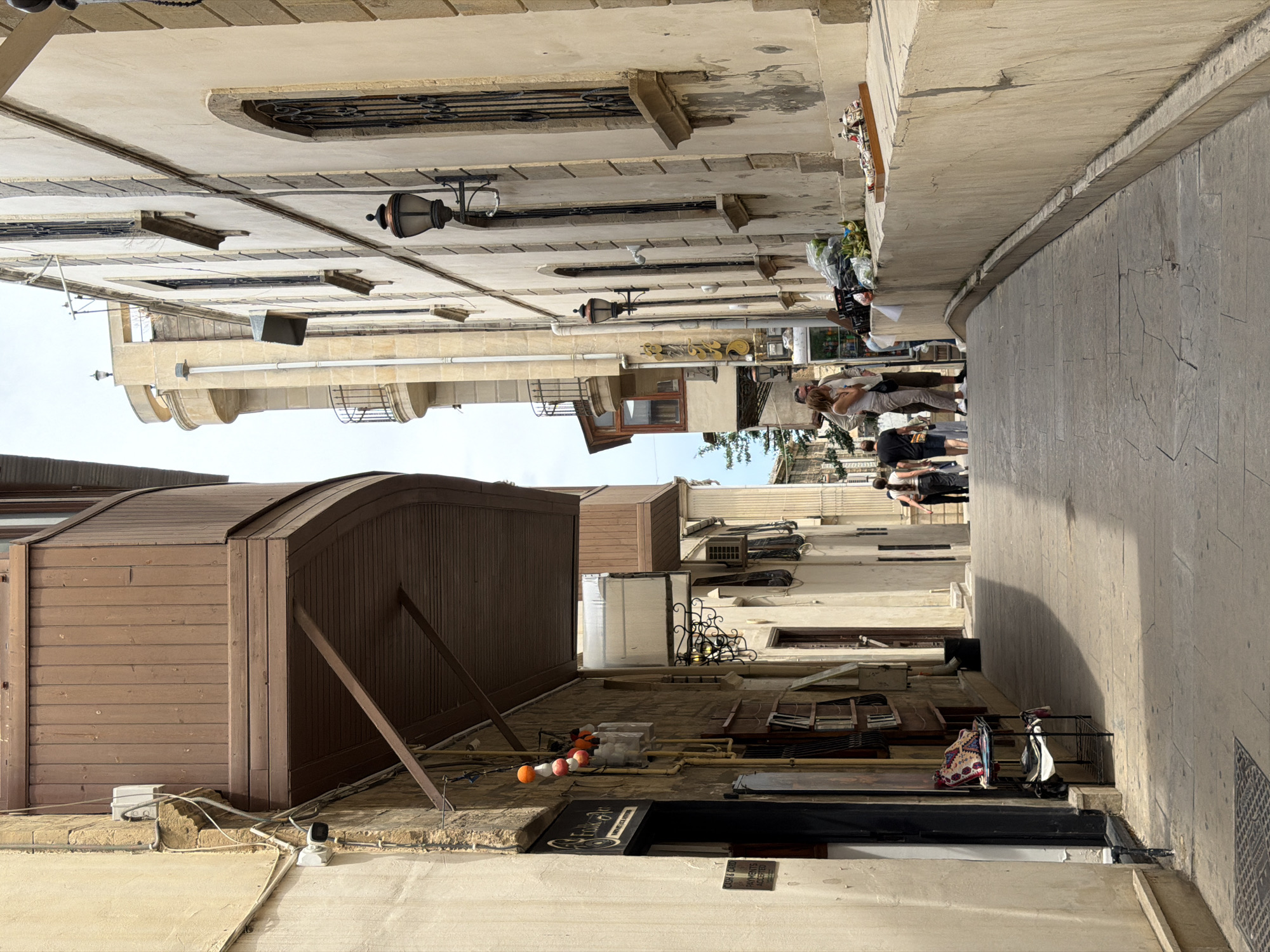 The image depicts a narrow street in the city of Sabail Raion, Azerbaijan. The street is paved with stone slabs and is flanked by tall, light-colored buildings. The architecture is traditional, with some buildings featuring wooden balconies and wrought-iron window grills. Sunlight casts shadows across the street, indicating it is daytime.
In the foreground, on the right side, a low, light-colored stone wall curves along the street. On top of this wall, a wooden tray holds several small, decorative items, possibly souvenirs. To the left of the wall, a pile of plastic-wrapped goods and some greenery are visible, suggesting a small stall or display.
Further down the street, several people are walking. In the middle ground, a couple stands near the stall area, facing each other, seemingly in conversation. The woman is wearing a white tank top and beige pants, and the man is wearing a light-colored shirt and darker pants. Behind them, other individuals are walking away from the camera.
The buildings on either side of the street are multi-story. On the left, a building has a dark doorway with a sign that reads "Folk Art." Above the doorway, a wooden balcony extends outward. Various pipes and electrical conduits are visible on the exterior walls of the buildings. On the right, the buildings have arched windows with ornate metalwork. A black, traditional-style street lamp is mounted on the building wall.
The sky is overcast and appears bright. The overall impression is of a historic urban environment, possibly a tourist area, with a mix of residential and commercial elements.