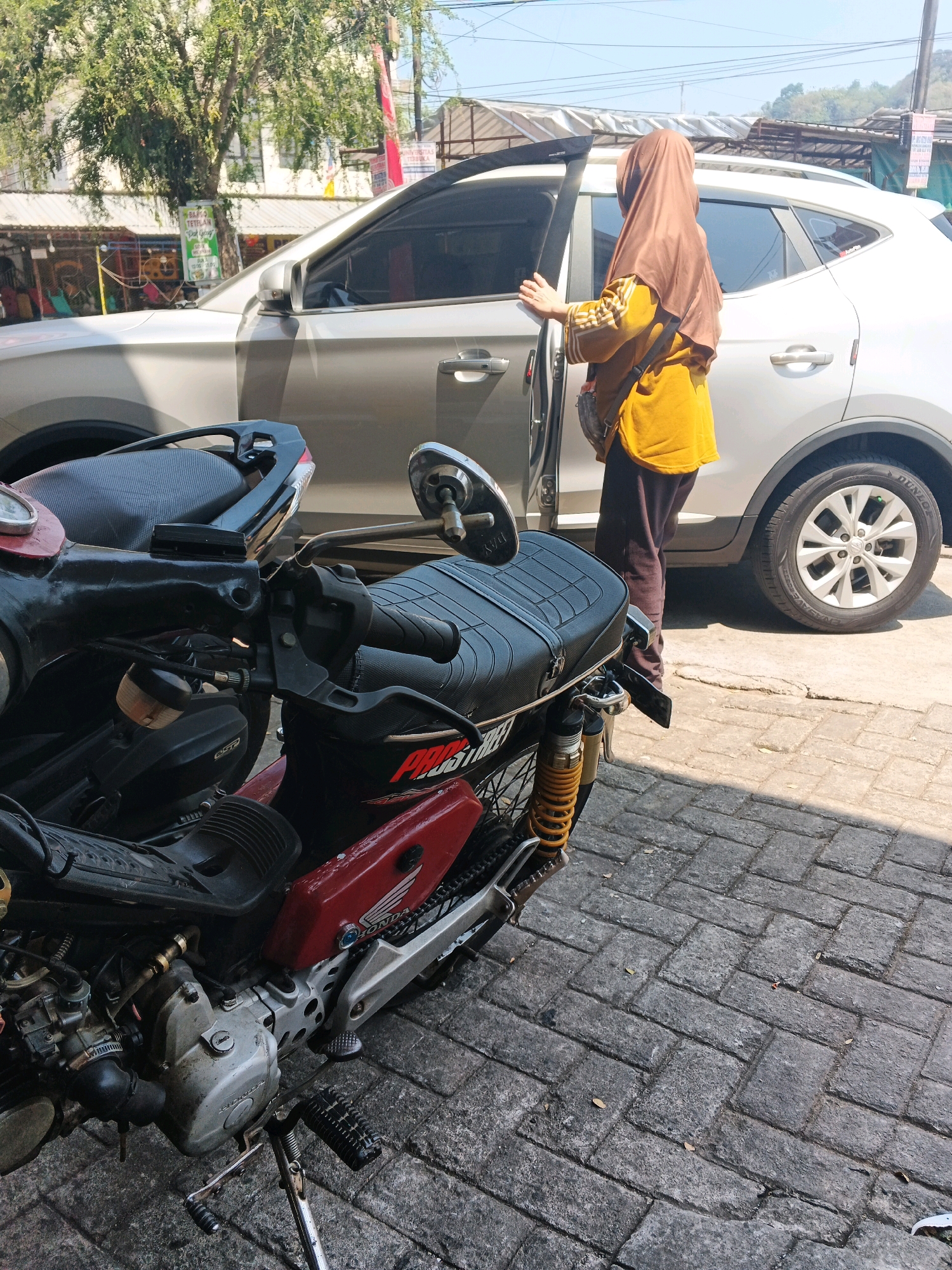 A person wearing a brown hijab and a yellow long-sleeved shirt stands next to an open car door in an outdoor urban setting, likely in Kalirejo, Indonesia. A motorcycle is parked in the foreground, partially obscuring the view of the person and the car. The car is a light-colored SUV, and the motorcycle is red and black. The ground is paved with interlocking bricks. Trees and buildings are visible in the background, suggesting a commercial or residential area. The scene appears to be well-lit by sunlight.
