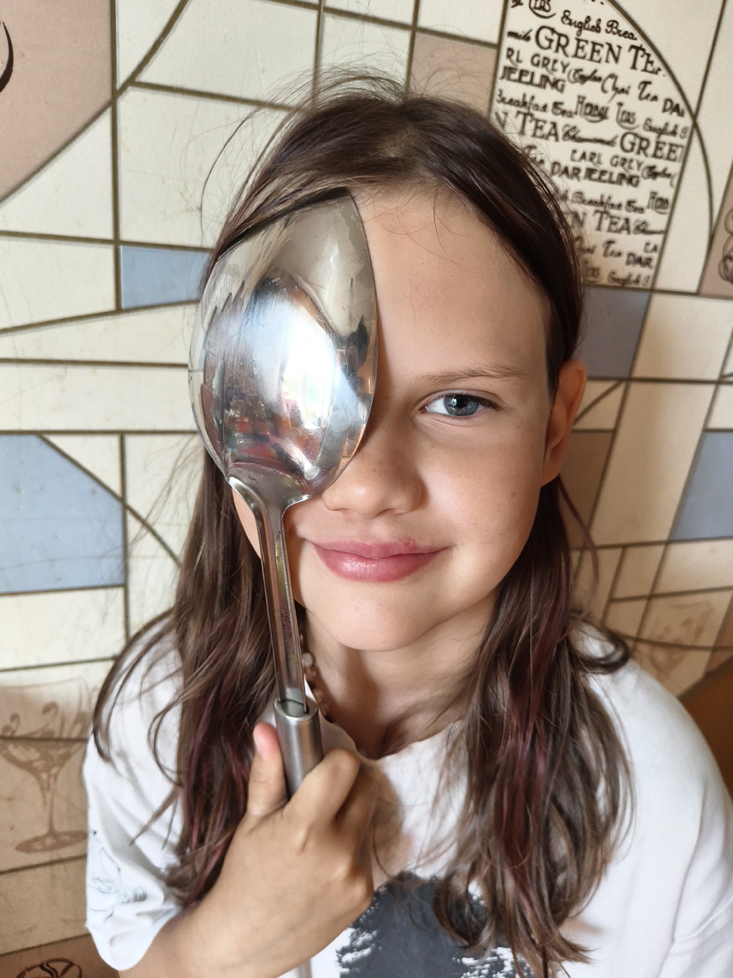 In an indoor setting, a young girl with long, brown hair, streaked with purple, covers her right eye with a large, metallic spoon. She looks directly at the camera with her visible blue eye and a slight smile. The background is a tiled wall with a repeating pattern of text related to tea and geometrical shapes. The lighting is even, suggesting an indoor environment.