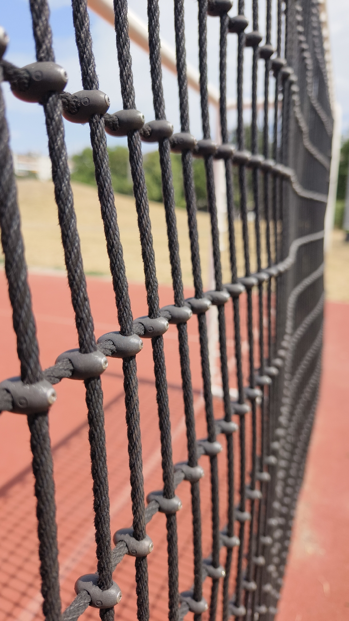 The image shows a close-up of a black rope net, likely part of a playground or sports facility, with small grey plastic fasteners securing the ropes. The ropes are arranged vertically and are in focus in the foreground, extending into the background where they become blurred. Behind the net, a red surface, possibly a sports court, is visible. Beyond that, there is a patch of dry grass and some greenery, with hints of buildings and a clear blue sky in the distance. There are no people or animals present in the image, and no discernible activities are taking place. The scene appears to be outdoors during daylight. The location is specified as Vienna, Austria, which suggests this is an outdoor public space within the city.