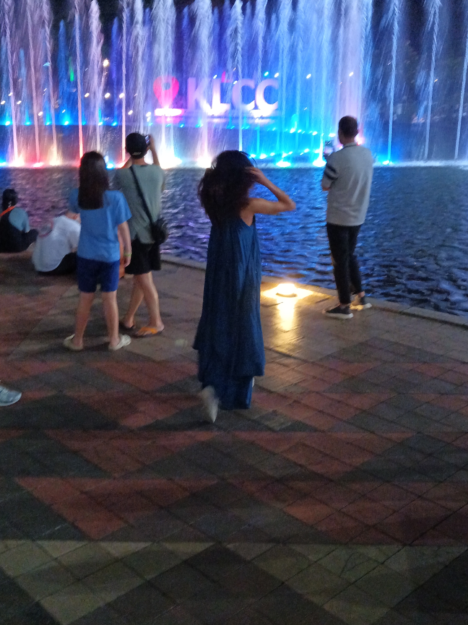 The image depicts an outdoor scene at night, likely a public space, with a prominent water fountain in the background. The fountain features numerous water jets illuminated by colorful lights, predominantly blue and pink. In the distance, large illuminated letters spelling "KLCC" are visible, indicating the location is near the Kuala Lumpur City Centre in Malaysia.
Several individuals are present in the foreground and middle ground. On the left, a person in a blue shirt and dark shorts stands facing away from the camera, holding a phone or camera. Next to them, a person in a gray t-shirt and dark pants is also facing away, holding what appears to be a phone to their face, possibly taking a picture. To the right, a person wearing a long, dark blue dress stands with their back to the camera, with their arms raised. Another individual, partially visible on the far left, is seated. In the middle ground, a person in a blue t-shirt and dark shorts is standing, also facing away.
The ground in the foreground is paved with a patterned brickwork, featuring diamond shapes in shades of brown, red, and gray. The overall atmosphere suggests a recreational or tourist setting, with people enjoying the illuminated fountain display.