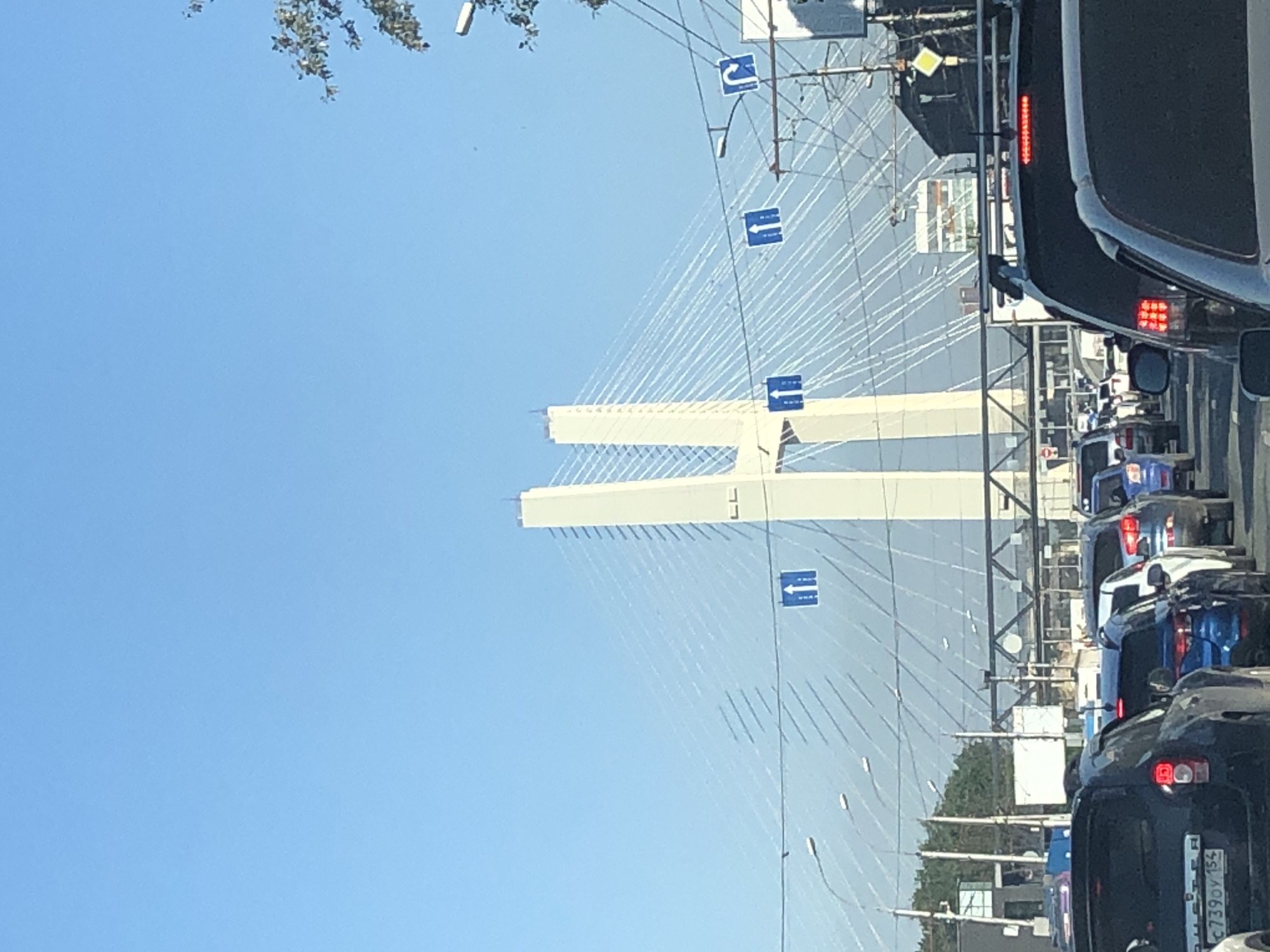 The image shows a traffic jam on a bright, clear day, with a large cable-stayed bridge dominating the background. The bridge has two tall, white pylons that are connected by numerous white cables. The pylons have the letters "LL" on them. Several blue road signs with white arrows are visible, indicating traffic direction. In the foreground, multiple cars are stopped in traffic, with some car details visible, including license plates. The environment appears to be urban, with some trees and buildings in the distance. The overall scene suggests a typical urban commute during daylight hours.