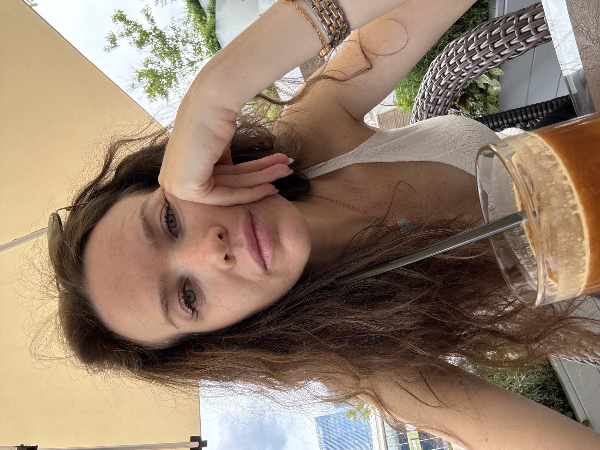 A close-up selfie captures a young woman seated at an outdoor café, looking directly at the camera. She has long, wavy brown hair, blue-green eyes, and light skin, and is wearing a light-colored ribbed top. Her expression is neutral to slightly pensive, with light pink lipstick. Her right hand is gently posed near her cheek. She wears a multi-toned bracelet on her right wrist and a delicate chain necklace with a small light blue pendant. Sunglasses are perched near her ear. A glass containing a brown beverage with a straw sits on the table in front of her.
The setting is a bright outdoor patio, shaded by a large tan umbrella or awning. In the background, green foliage, a wicker chair, and modern urban buildings with glass facades under a partly cloudy sky are visible, indicating a bustling city environment. The bright natural light suggests it is daytime, likely late morning or afternoon. The scene conveys a relaxed moment in an urban outdoor setting, consistent with the provided context of Moscow, Russia. No visible text is legible.