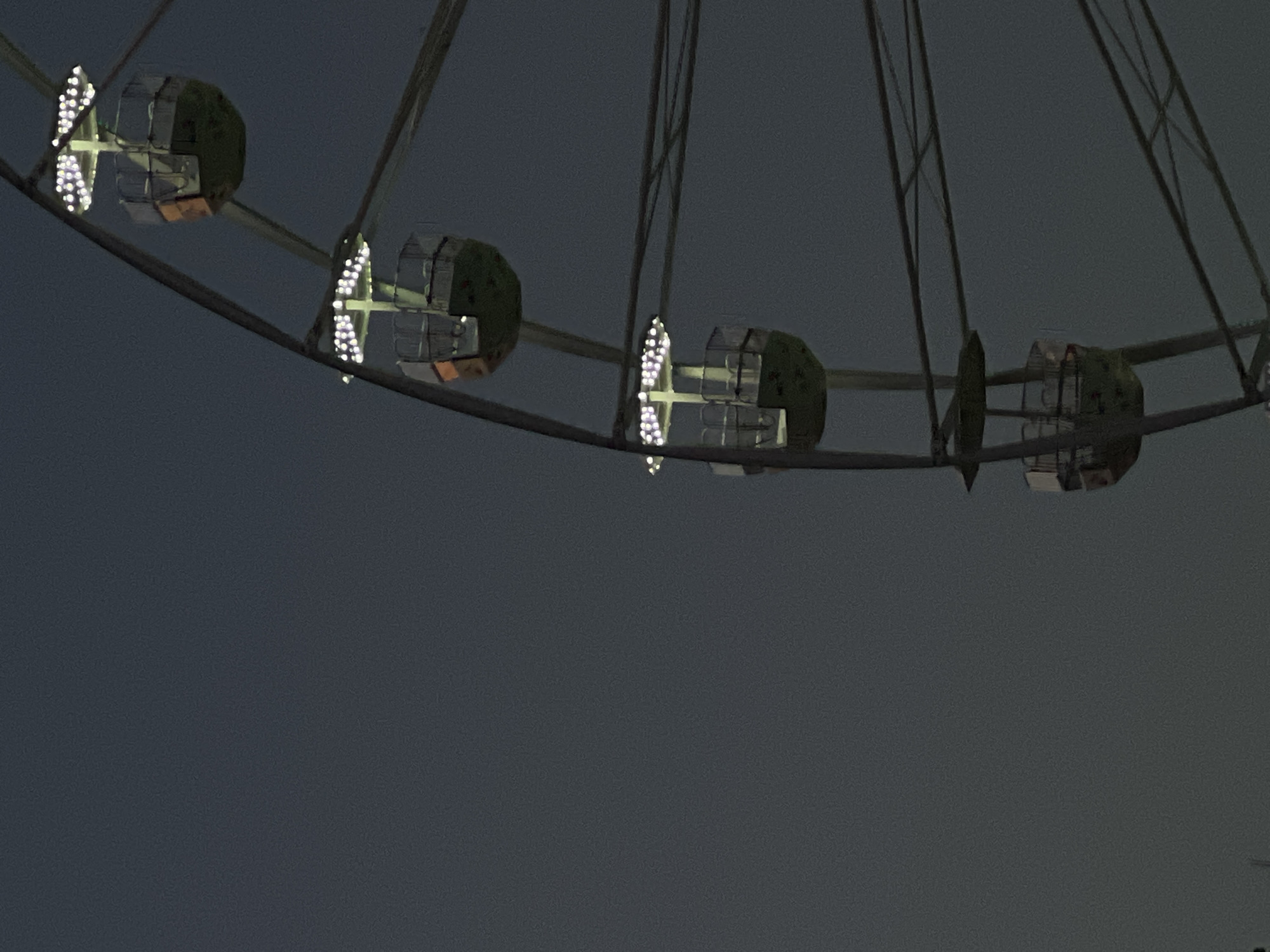 The image is a low-angle, close-up shot of a Ferris wheel against a dark, overcast sky. Several gondolas are visible, each illuminated by strings of small, white lights. The gondolas are suspended from the wheel's structure by metal arms. The Ferris wheel appears to be operational, though no people are clearly visible within the gondolas. The overall atmosphere is subdued due to the low light conditions and the dark sky. The scene does not contain any specific architectural or geographical features that would allow for precise identification of its location within Sofia, Bulgaria.