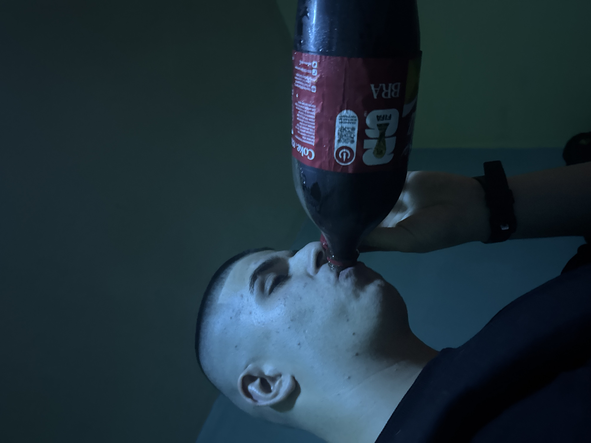 A man is drinking from a large bottle of Coca-Cola. The bottle's label indicates it is the FIFA World Cup Brazil edition. The man is fair-skinned with a close-cropped haircut. He is wearing a black shirt. The scene is dimly lit. The location is listed as Unknown, Unknown.