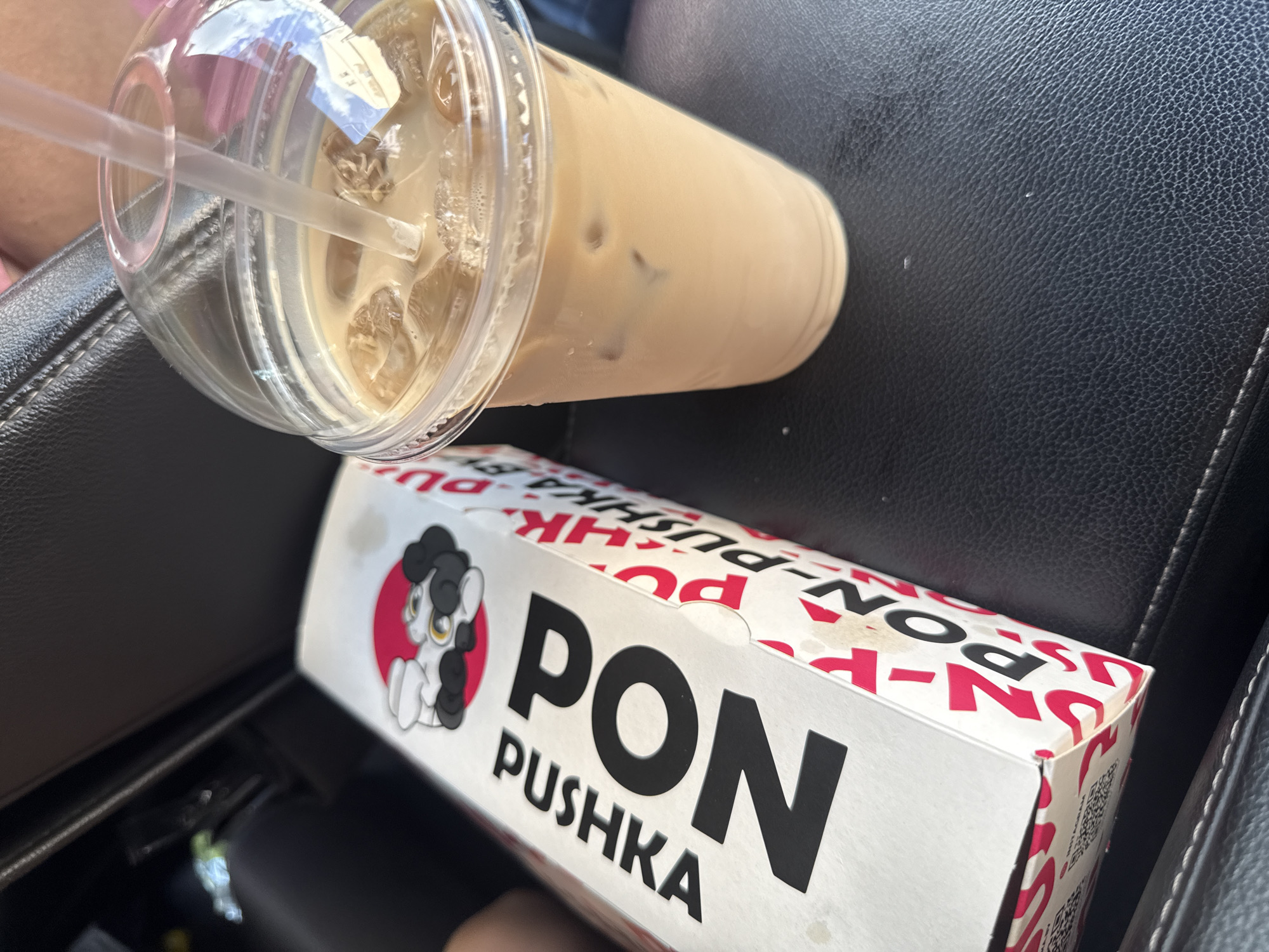 A clear plastic cup filled with an iced coffee drink and a straw is positioned diagonally in the upper left corner of the image. The drink is a light brown color with ice cubes visible. Below the cup is a white cardboard box with red and black lettering and graphics. The box has a cartoon drawing of a white pony with black hair and a pink circle behind it on the left side. Large black text on the box reads "PON" and below that, smaller black text reads "PUSHKА." Red lettering, which appears to be repeating "PON-PUSHKA," is visible on the box as well. The background appears to be a dark, textured surface, possibly car upholstery. The lighting suggests it is daytime. The overall scene appears to be inside a vehicle, possibly in Mahilyow, Belarus, given the location context. There is no discernible activity or event taking place beyond the items being present.