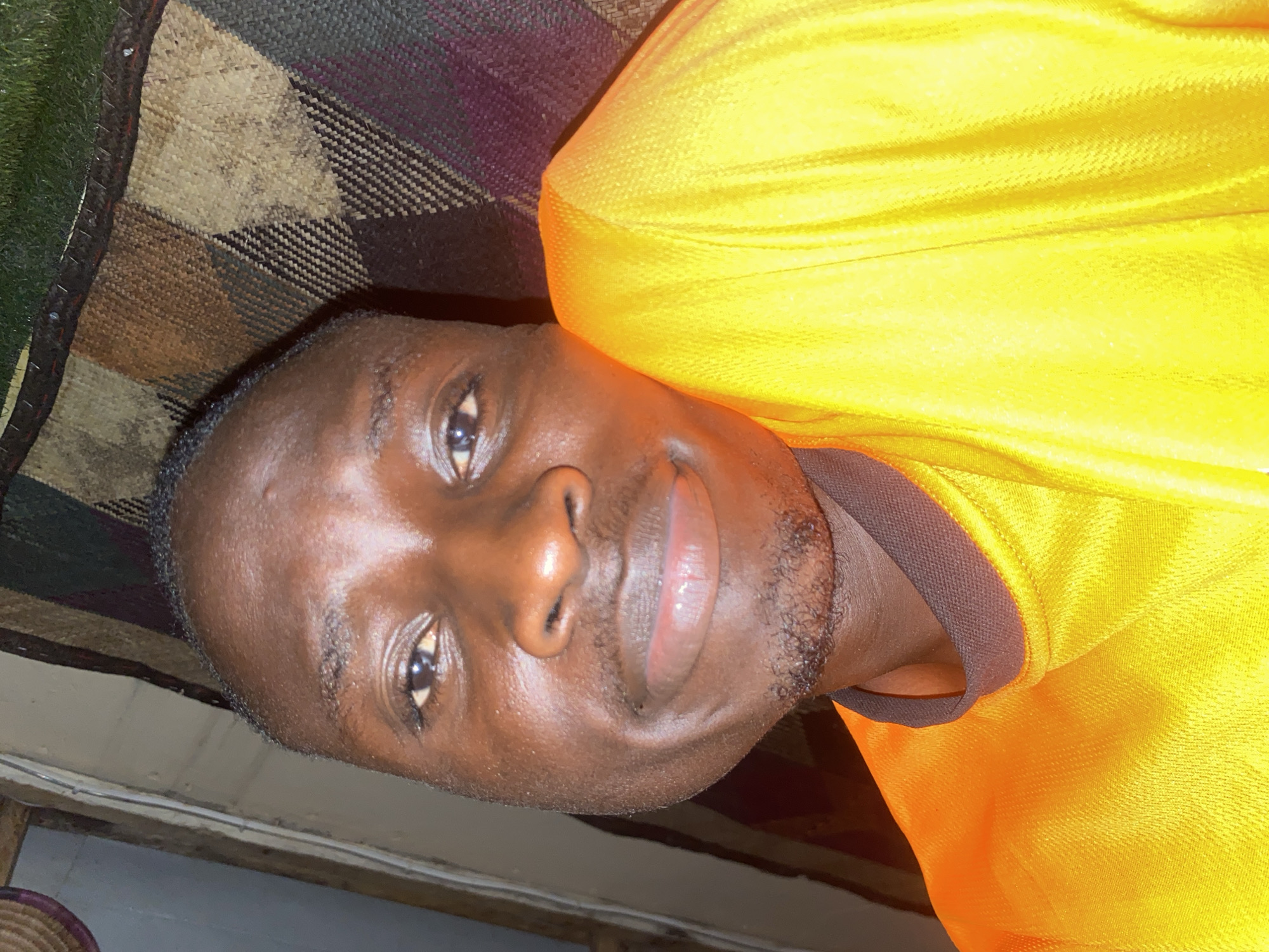 This media file presents a portrait of a man, appearing to be of African descent, looking directly at the camera with a calm and open expression, possibly a slight smile. He has dark skin, short dark hair, and neatly trimmed facial hair, including a goatee and stubble. He is wearing a bright yellow short-sleeved shirt with a dark brown collar or trim visible around his neck and shoulder.
The man appears to be reclining or lying down, with his head tilted. In the background on his left side, a patterned woven mat or blanket is visible, featuring a design with shades of brown, beige, green, and dark purple/black. The background on his right side is darker and less defined, possibly a floor or lower wall, with a horizontal wooden element visible. A small portion of a woven basket can be seen in the bottom left corner of the frame.
The lighting is relatively bright and appears to be direct, casting highlights on his skin and reflecting in his eyes. There is no visible text in the image. The setting, characterized by the woven mat and basket, suggests an indoor or sheltered environment, consistent with the provided location context of Barden Yana, Nigeria. The time of day is not discernible from the image.
