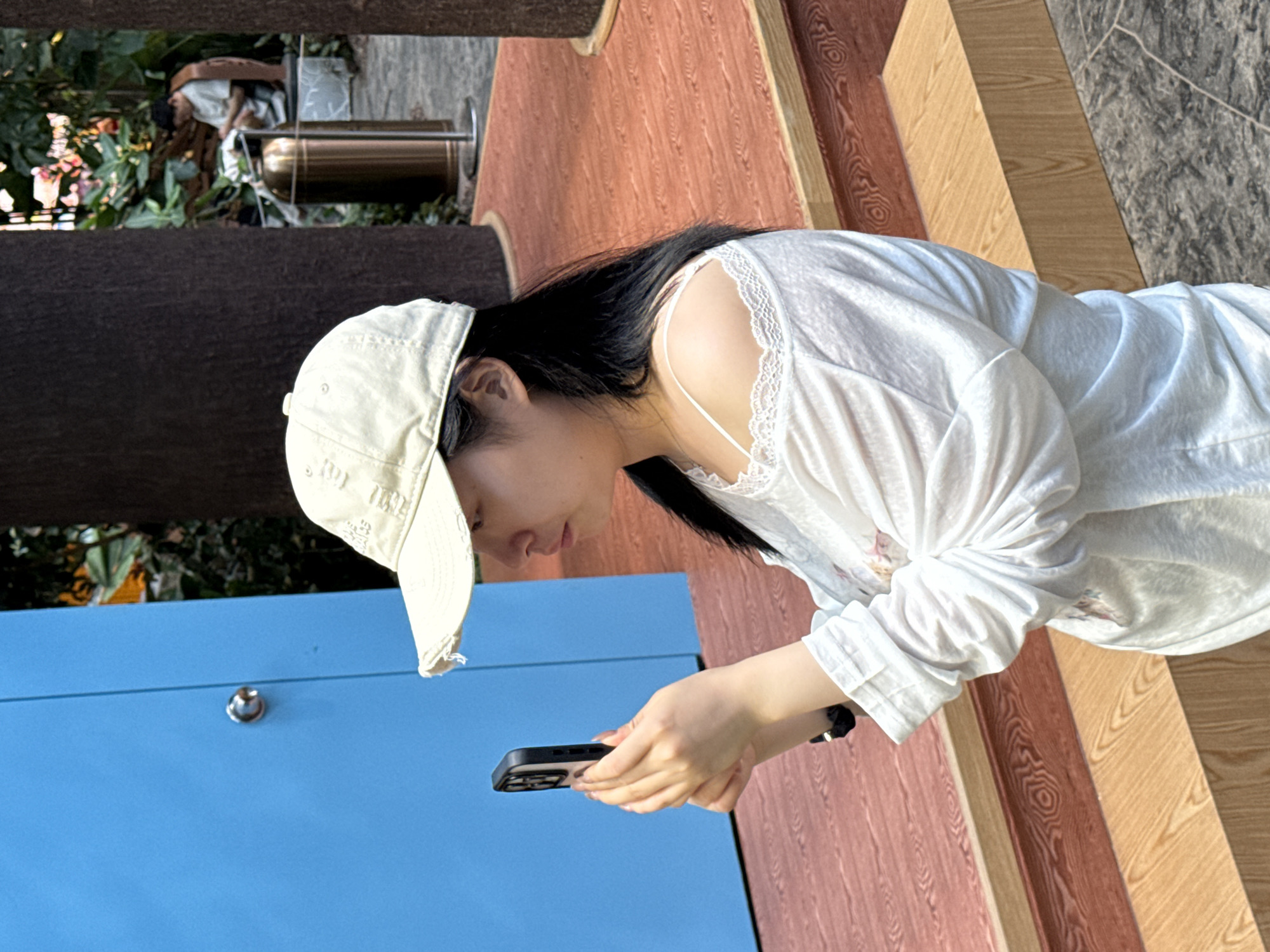 A person wearing a white cap and a white, long-sleeved shirt is looking at a cell phone held in their hands. The background includes a blue cabinet with a small, round metal knob, outdoor greenery, a metal pole structure, and a floor surface composed of planks with different wood-grain patterns. The location may be Liangjiang, China.