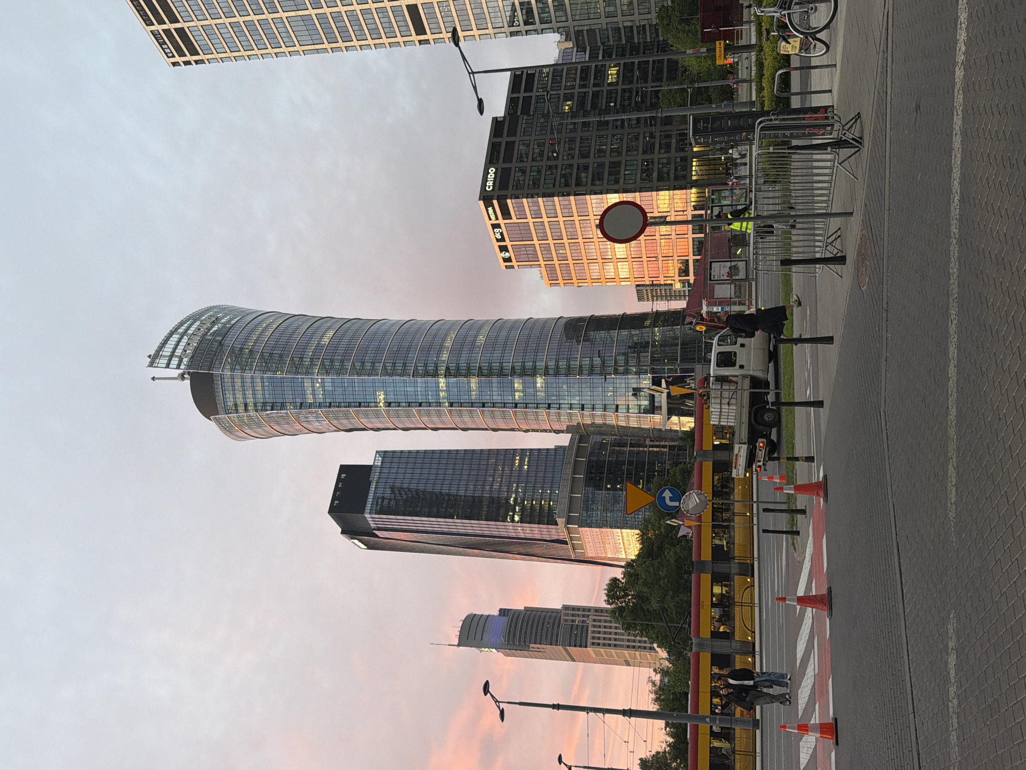 The image depicts a cityscape, likely at dusk or dawn given the pink-tinged sky. Dominating the scene are several tall, modern glass buildings, including a uniquely curved skyscraper. Street-level details include a crosswalk marked with orange cones, a yellow tram or bus, and a few pedestrians. A white truck or van is parked near the sidewalk. Street signs and construction barriers are also visible. The location is unknown, but based on architectural styles and streetscape, it could be a major city in Europe.