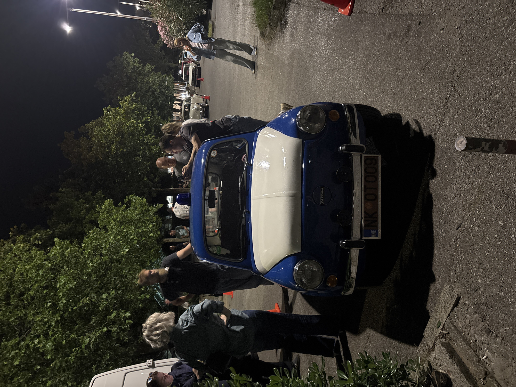 The image shows a dark, nighttime scene featuring a blue and white Zastava 750 car parked on an asphalt surface, likely a street or parking lot. Several people surround the car, some appearing to examine it closely. Others are standing nearby, seemingly part of the gathering. A few additional cars are visible further down the road. The license plate of the Zastava shows it is from Montenegro, specifically the "NK" designation, indicating the Nikšić municipality. Given that Nikšić is a common destination from Podgorica, it is plausible the scene is in or near Podgorica, Montenegro. There are trees and some greenery on the left side. Streetlights illuminate the area. An orange traffic cone is to the left and above the car.
