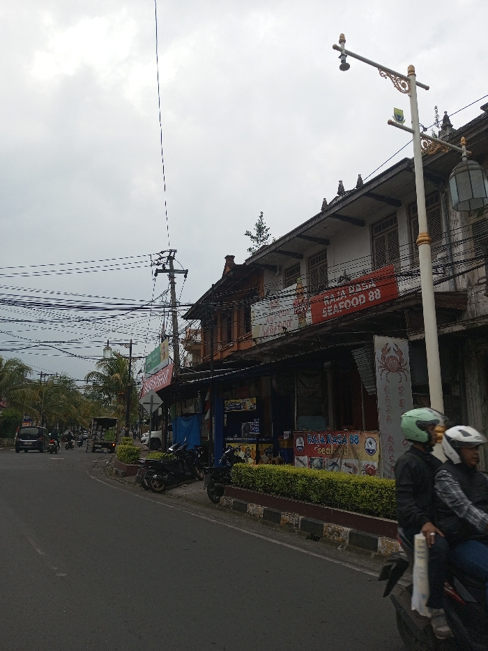 A street scene showcases businesses under a cloudy sky. Motorbikes are parked along the curb and a motorcycle with two riders passes by. A sign for "RAJA RASA SEAFOOD 88" is visible on a building. Power lines crisscross the sky, and a decorative street lamp stands tall. The buildings are older with ornate details. Green hedges line the curb. Traffic moves along the road.