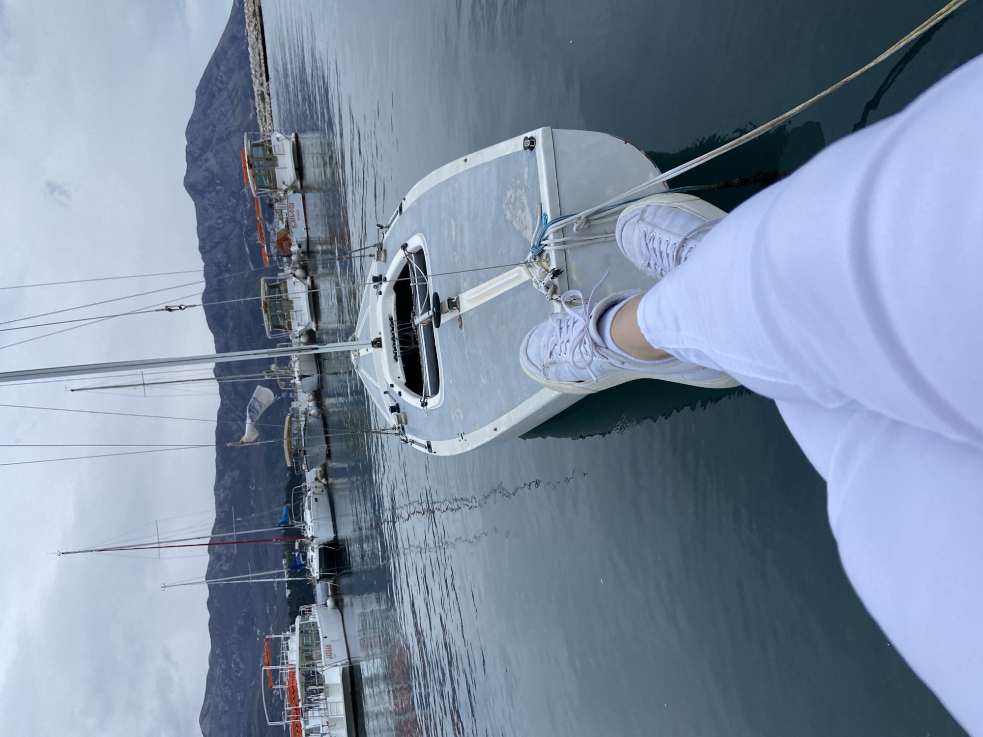 A person wearing white sneakers and white pants is sitting on a sailboat docked in a marina. The boat is gray and white. There are other boats docked nearby. In the background is a mountain range and a cloudy sky. The marina is located in Budva, Montenegro.